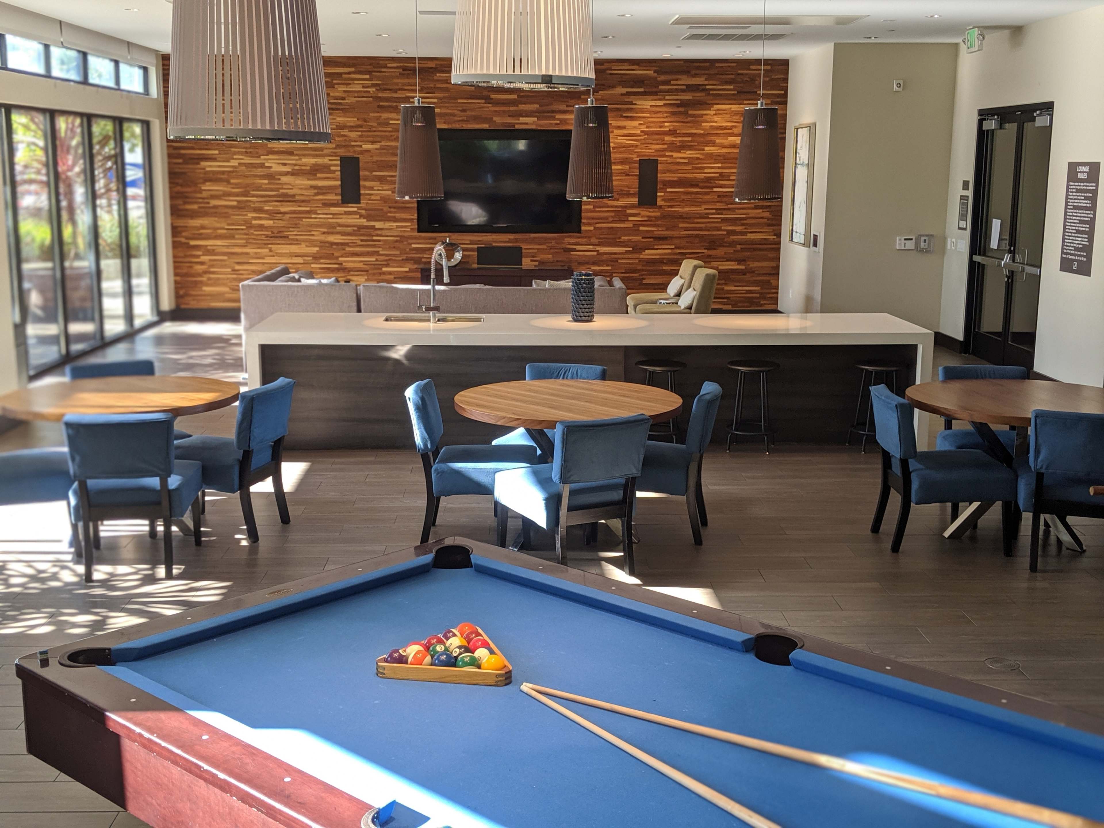 A modern lounge area featuring a billiard table in the foreground, round tables with blue chairs, and a stone wall with a television in the background.