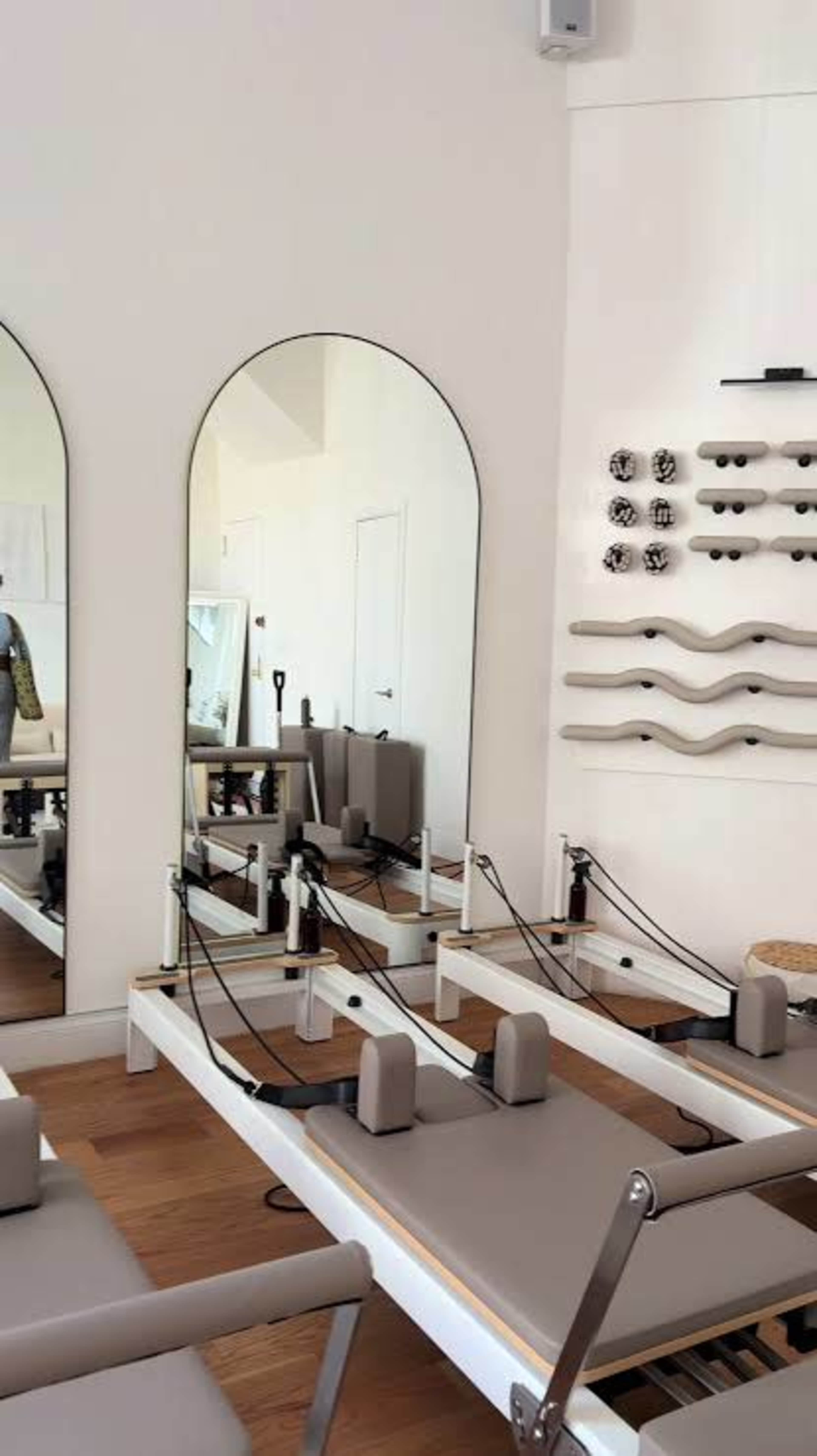 The image shows a modern Pilates studio featuring reformer machines, mirrors, and exercise equipment mounted on a wall.