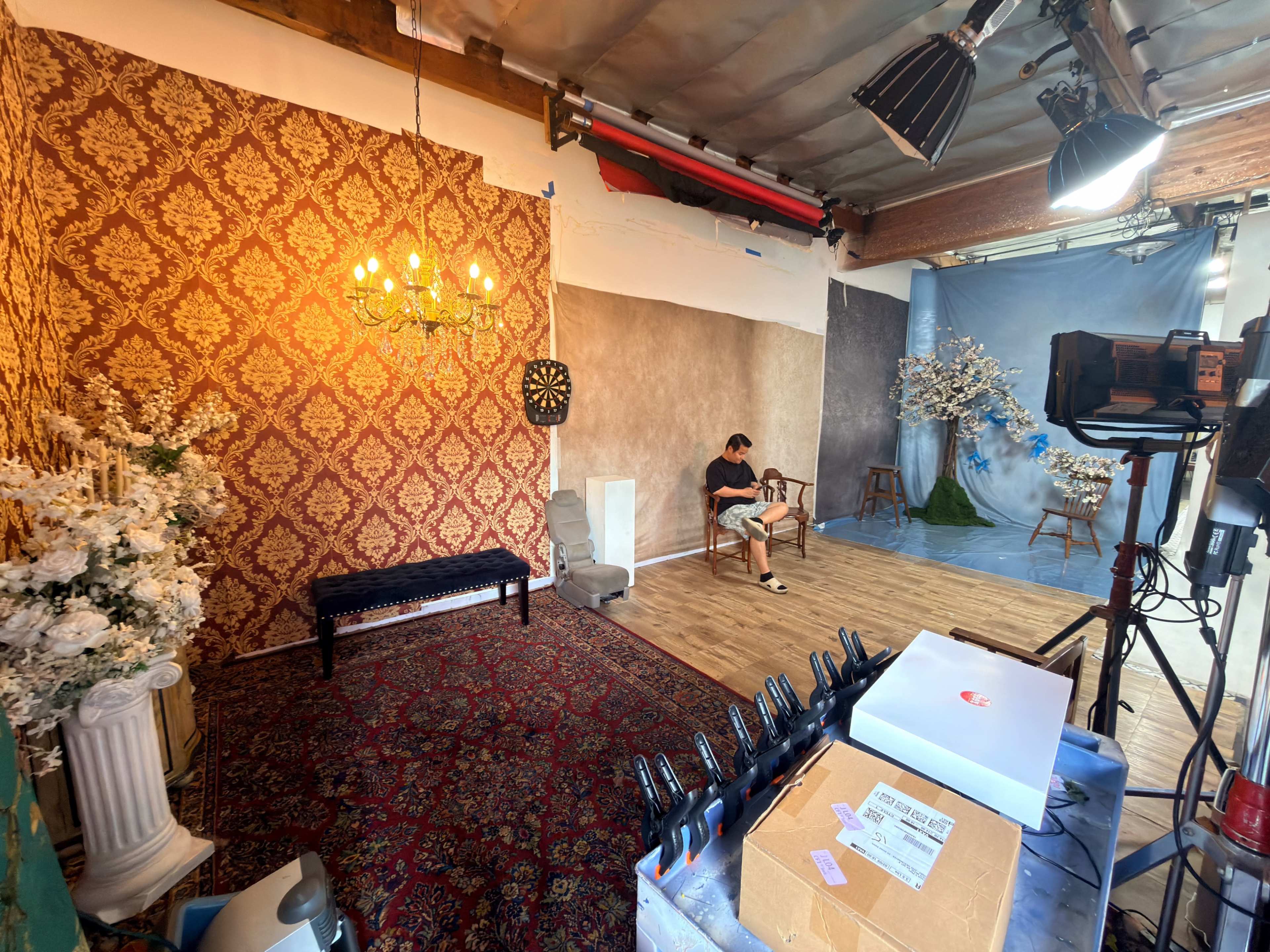 The image shows a photography studio with various backdrops, a chandelier, a dartboard, and a person sitting on a bench.