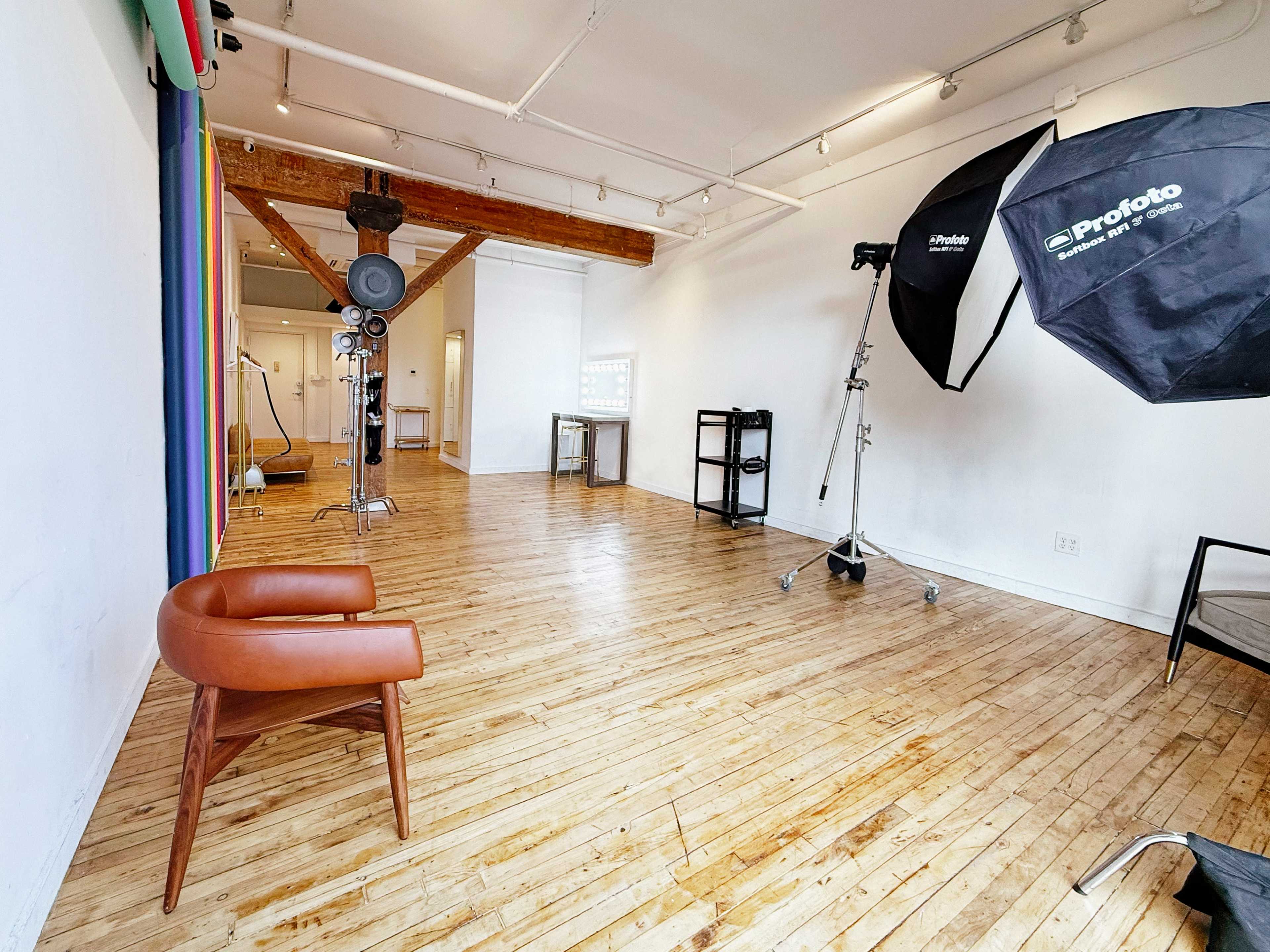 The image shows a spacious photo studio with wooden floors and minimal decor, featuring a chair, lighting equipment, and a backdrop on one wall.