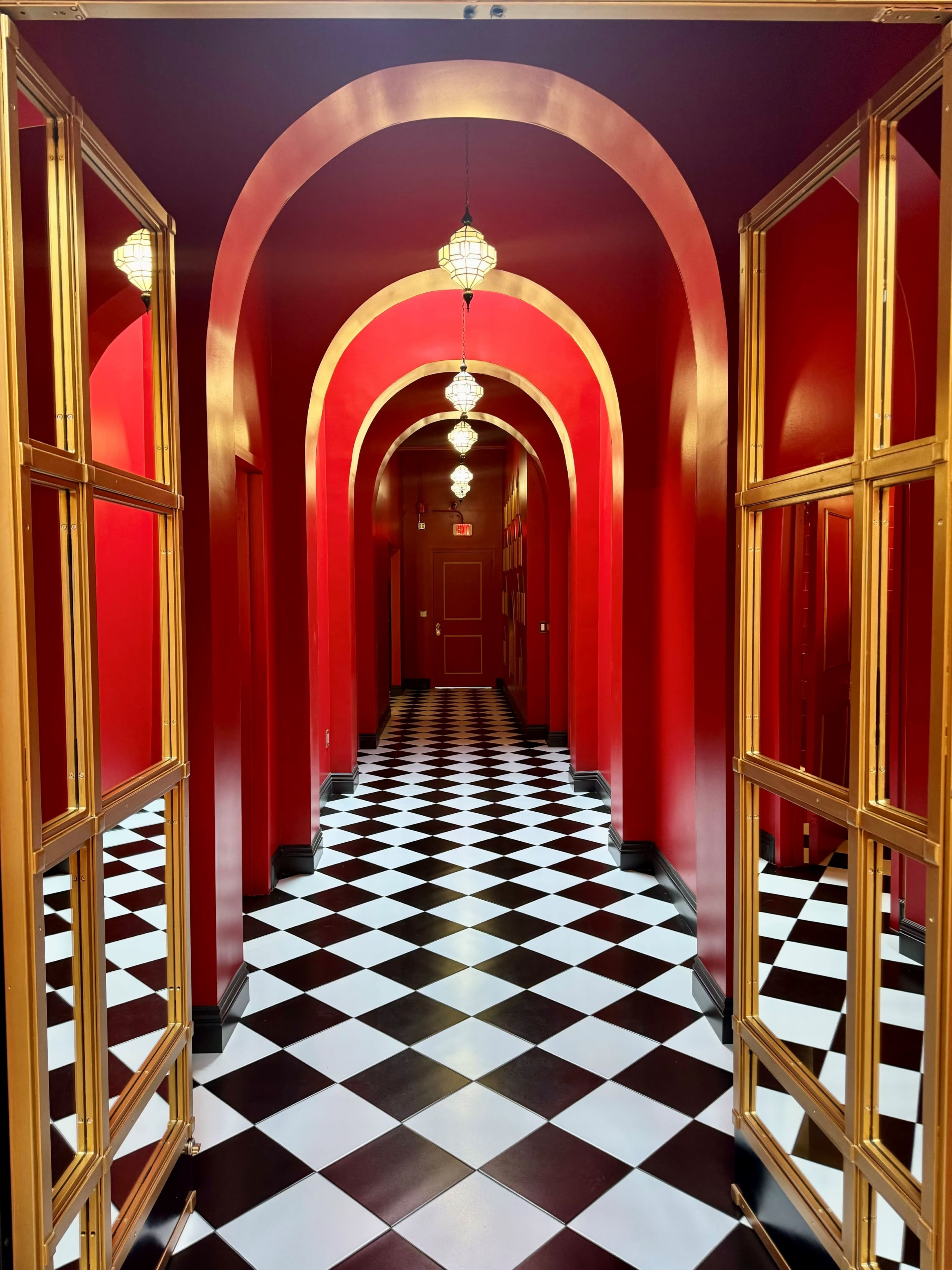 A corridor with red walls and arches, featuring a black and white checkered floor and golden-framed mirrors leading into the distance.