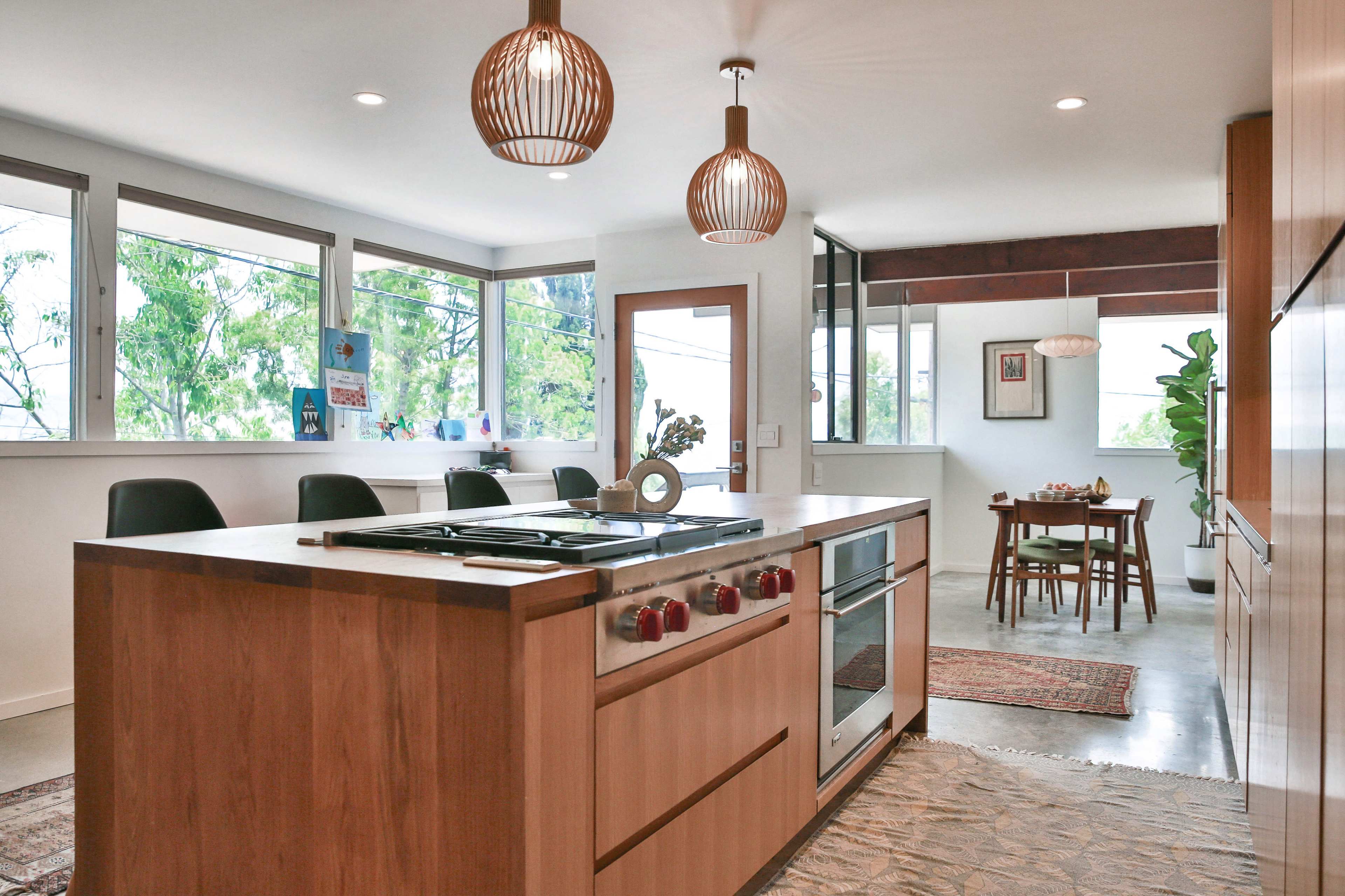 The image shows a modern kitchen with wooden cabinetry, a central island featuring a stove, and a dining area with a table and chairs in the background.