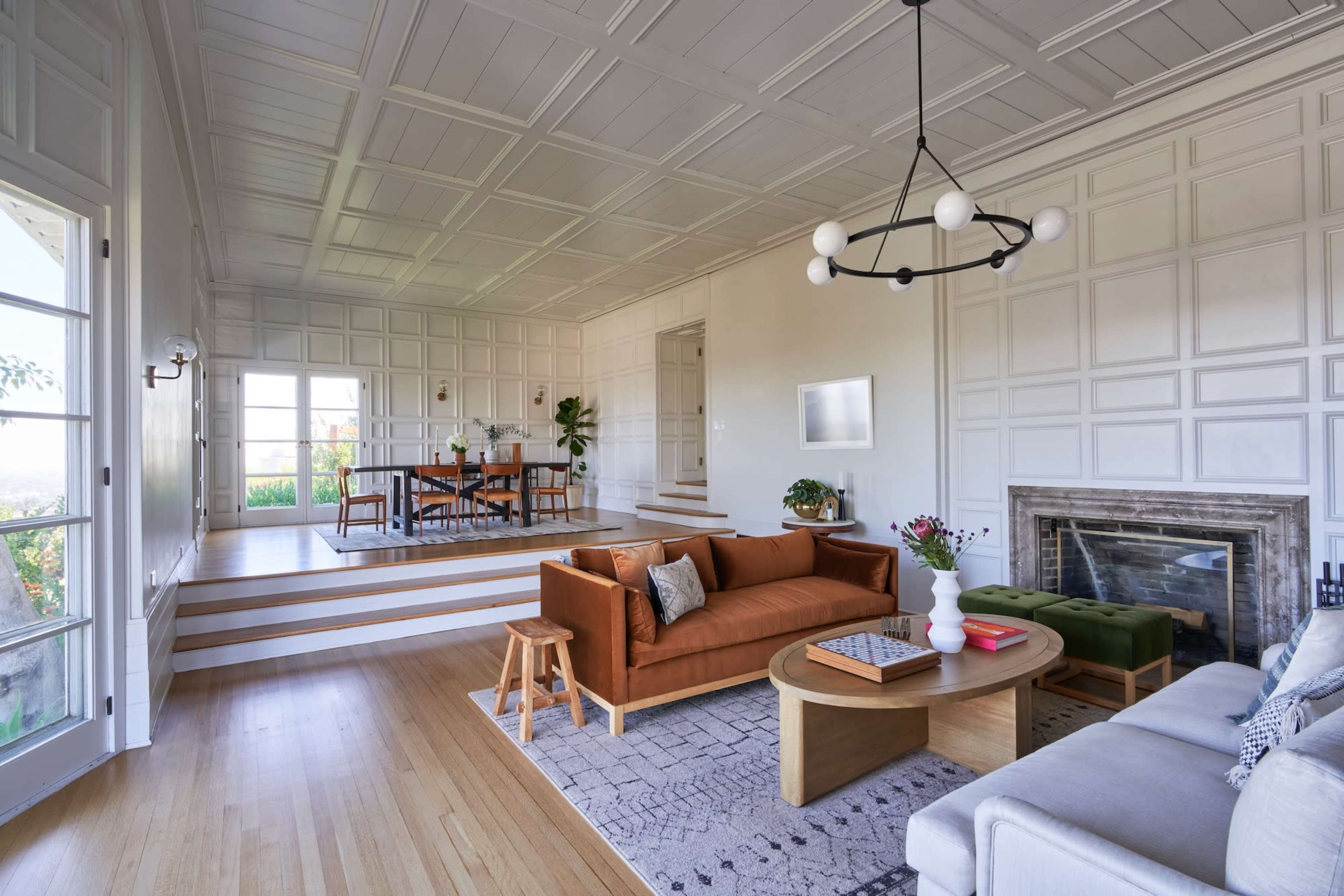 The image shows a spacious living room with a sectional sofa, a round coffee table, and a dining area in the background featuring a wooden table and chairs.