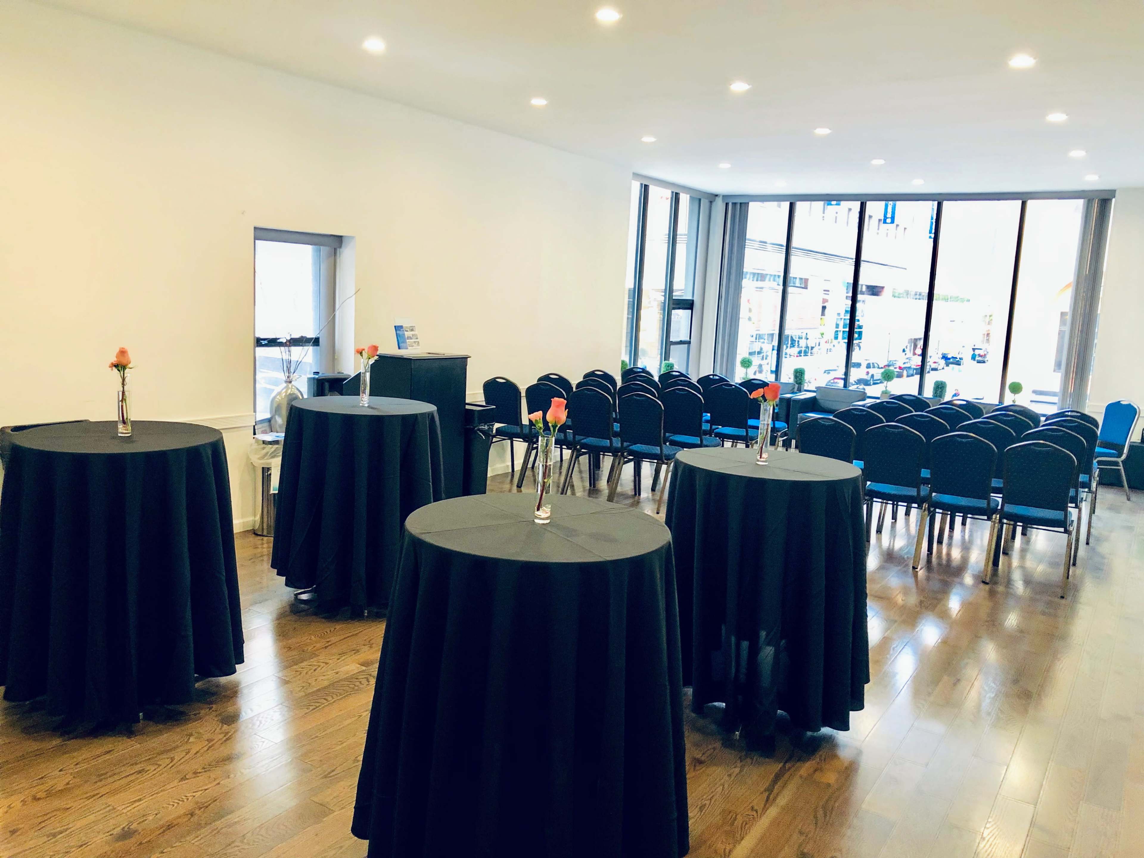 A spacious event room features several black round tables with floral centerpieces and a row of chairs arranged against a large window.