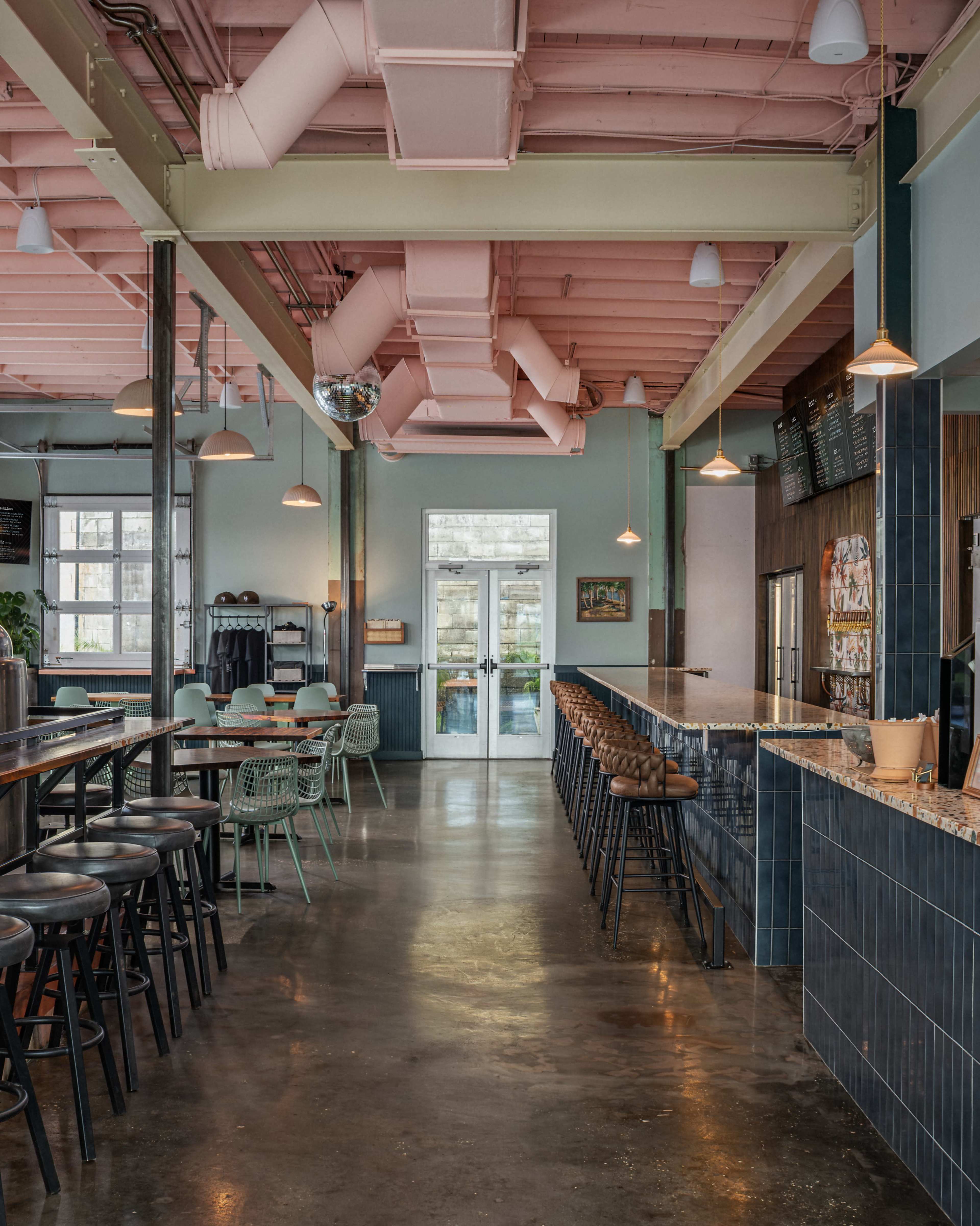 The image shows a modern, spacious café with a long bar, colorful chairs, and large windows letting in natural light.