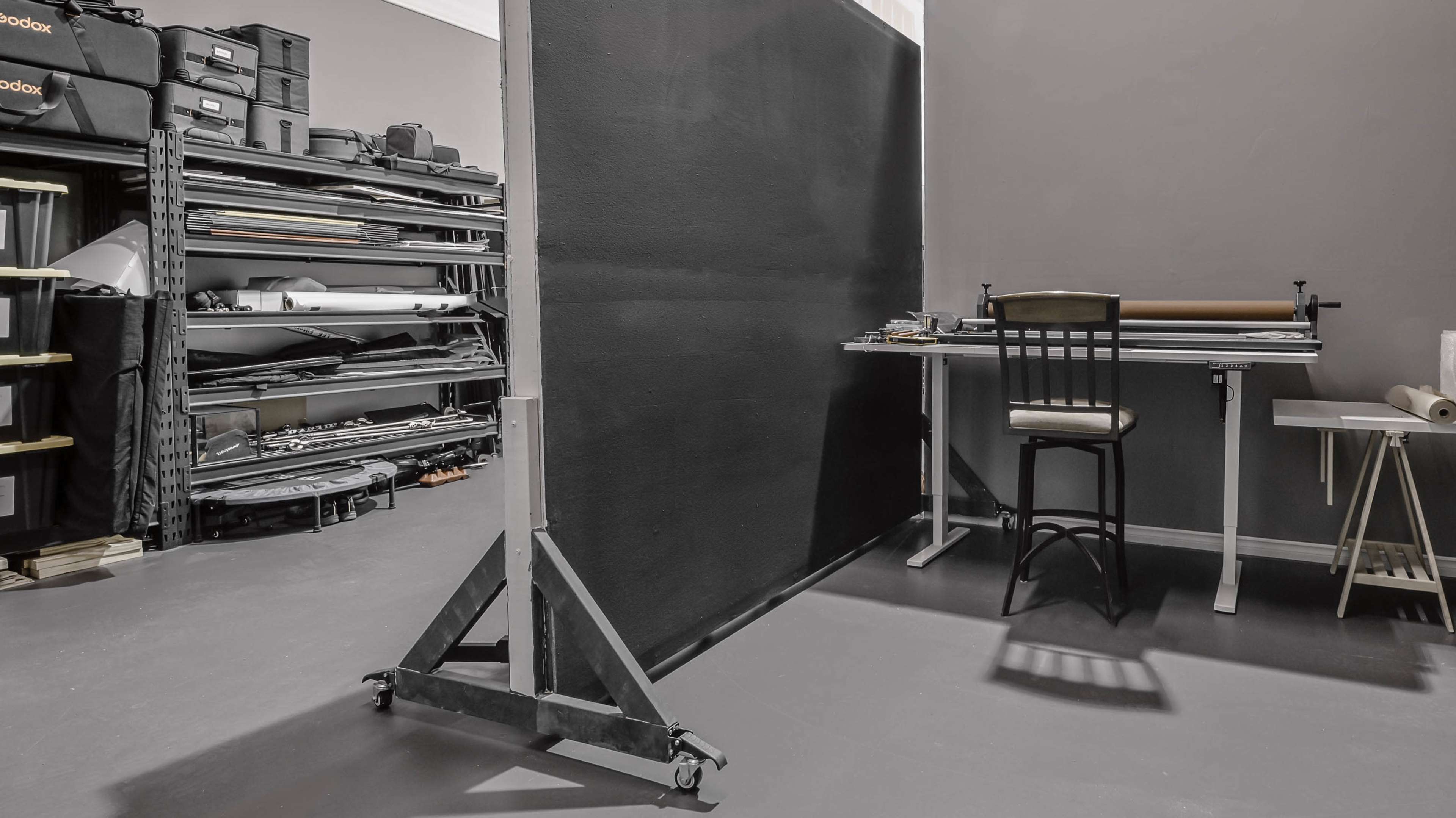 The image shows a work area with a tall black partition, a table with a chair, and shelves filled with various storage boxes and equipment.