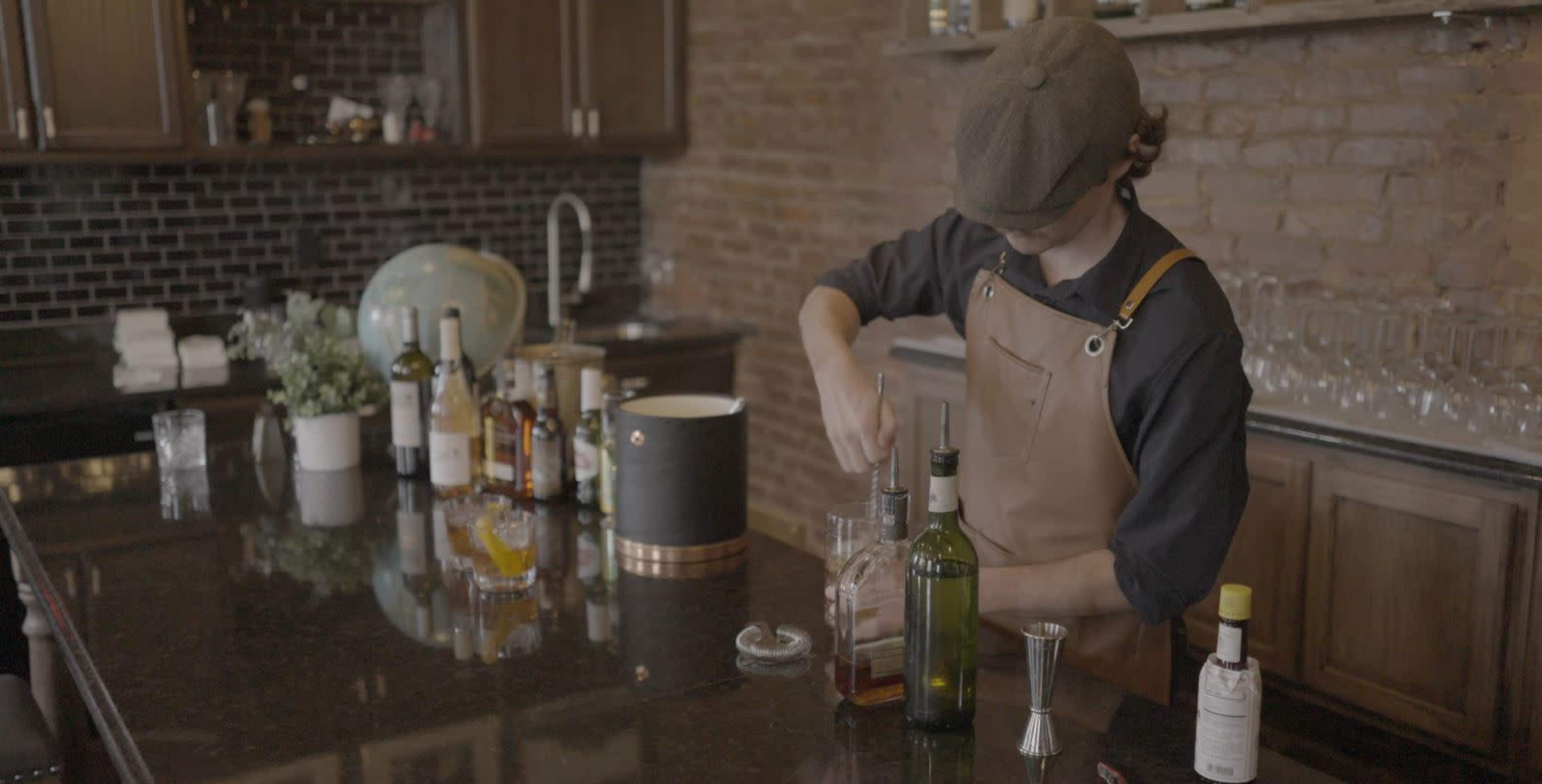 A bartender in a brown apron mixes a drink at a dark countertop filled with various bottles and bar tools.