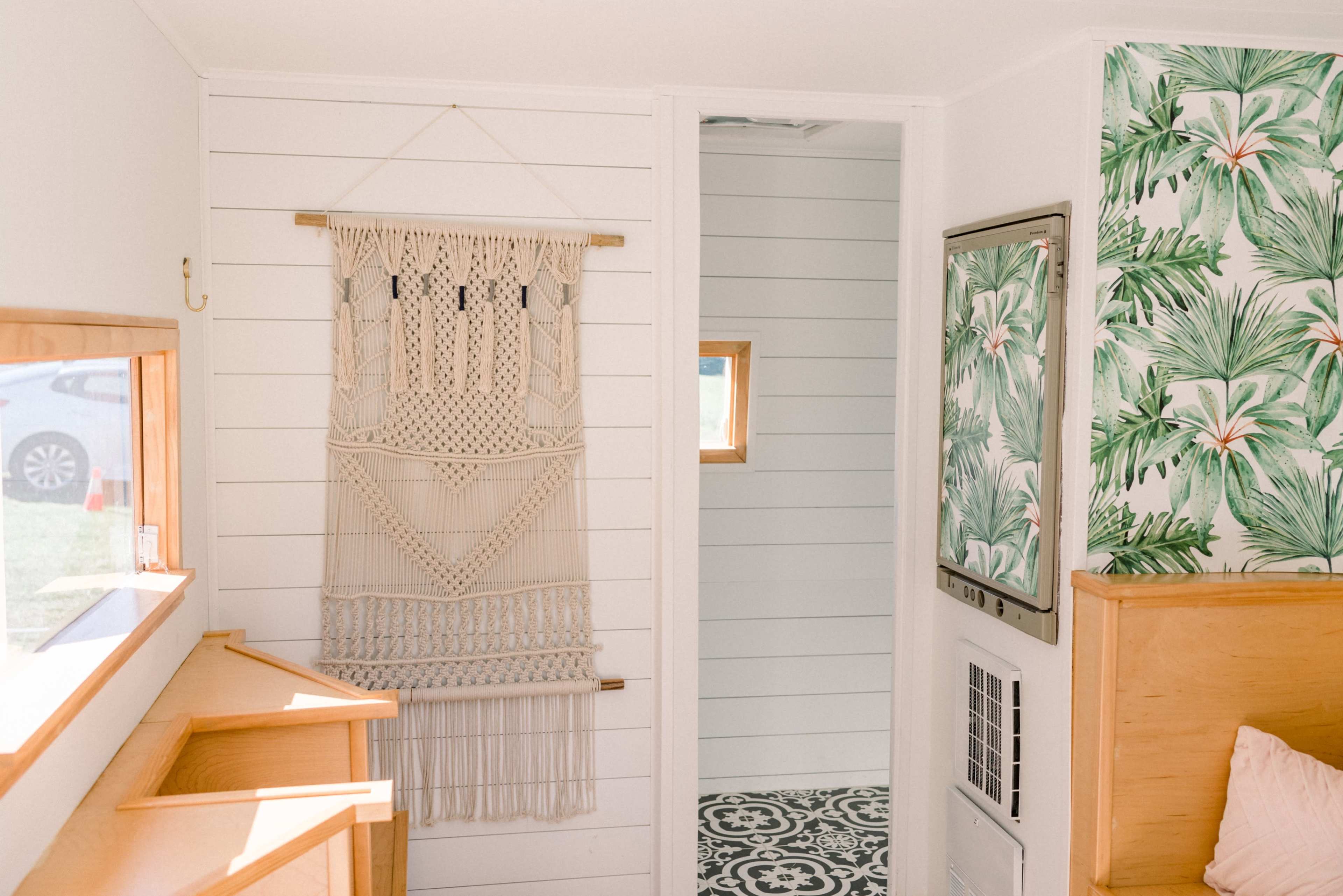 A bright interior featuring a macramé wall hanging, a mirrored frame, and a tropical-themed wallpaper with green foliage.
