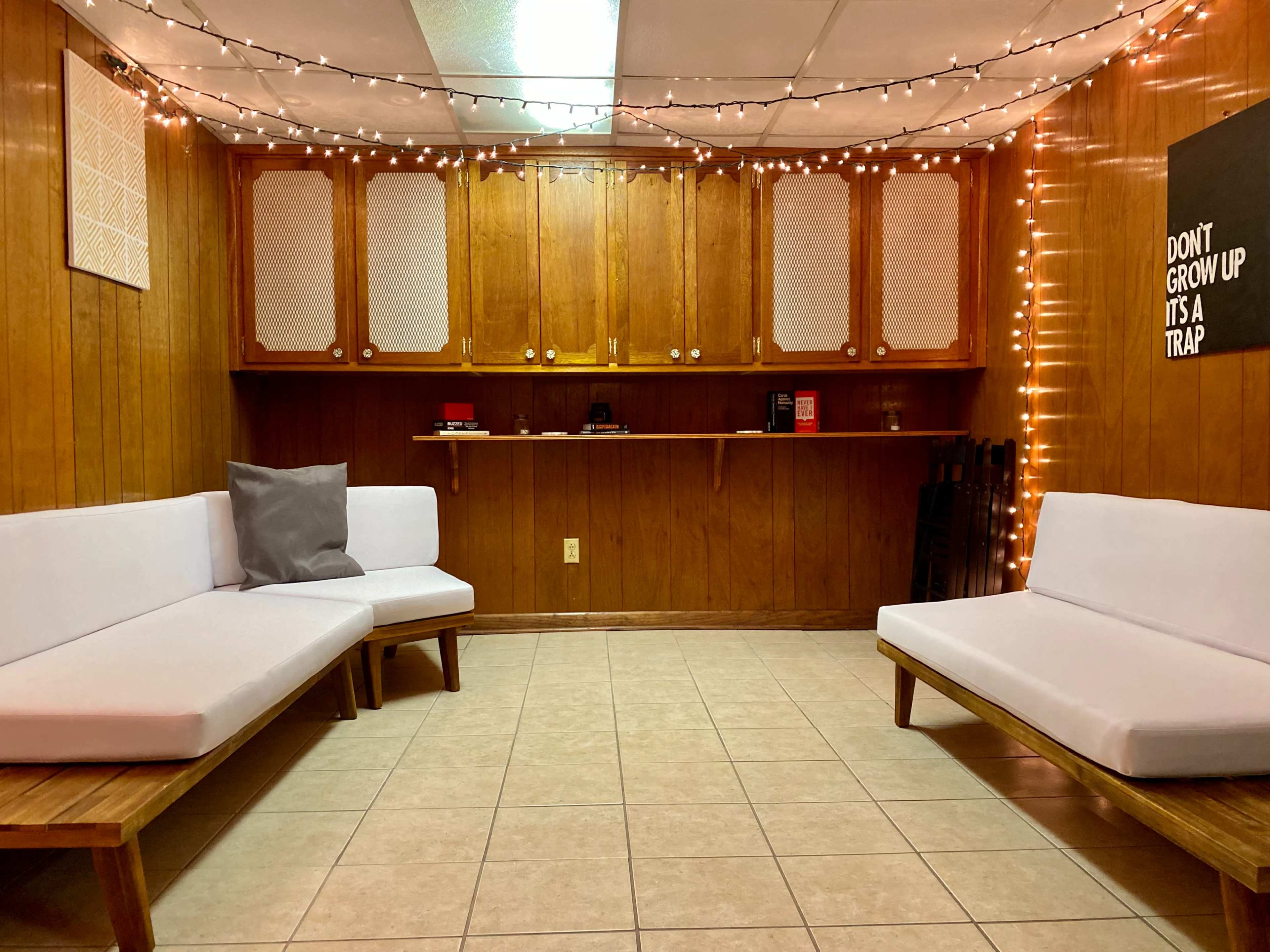 A room with wooden paneling, two white couches, a tiled floor, and a bar area illuminated by string lights.
