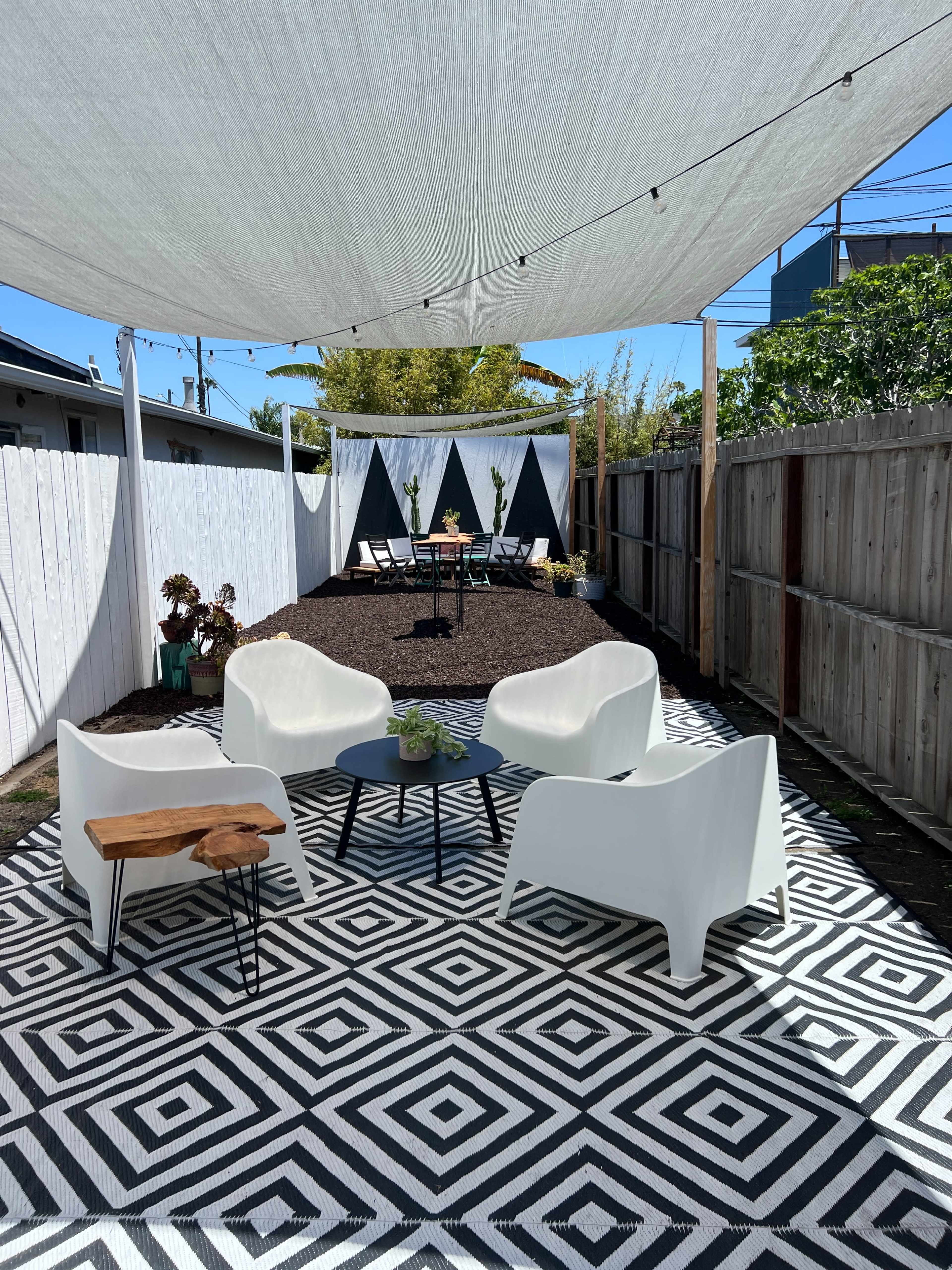 A patio area features white chairs arranged around a circular table on a geometric-patterned rug, with a shaded section and a backdrop of decorative fabric triangles.