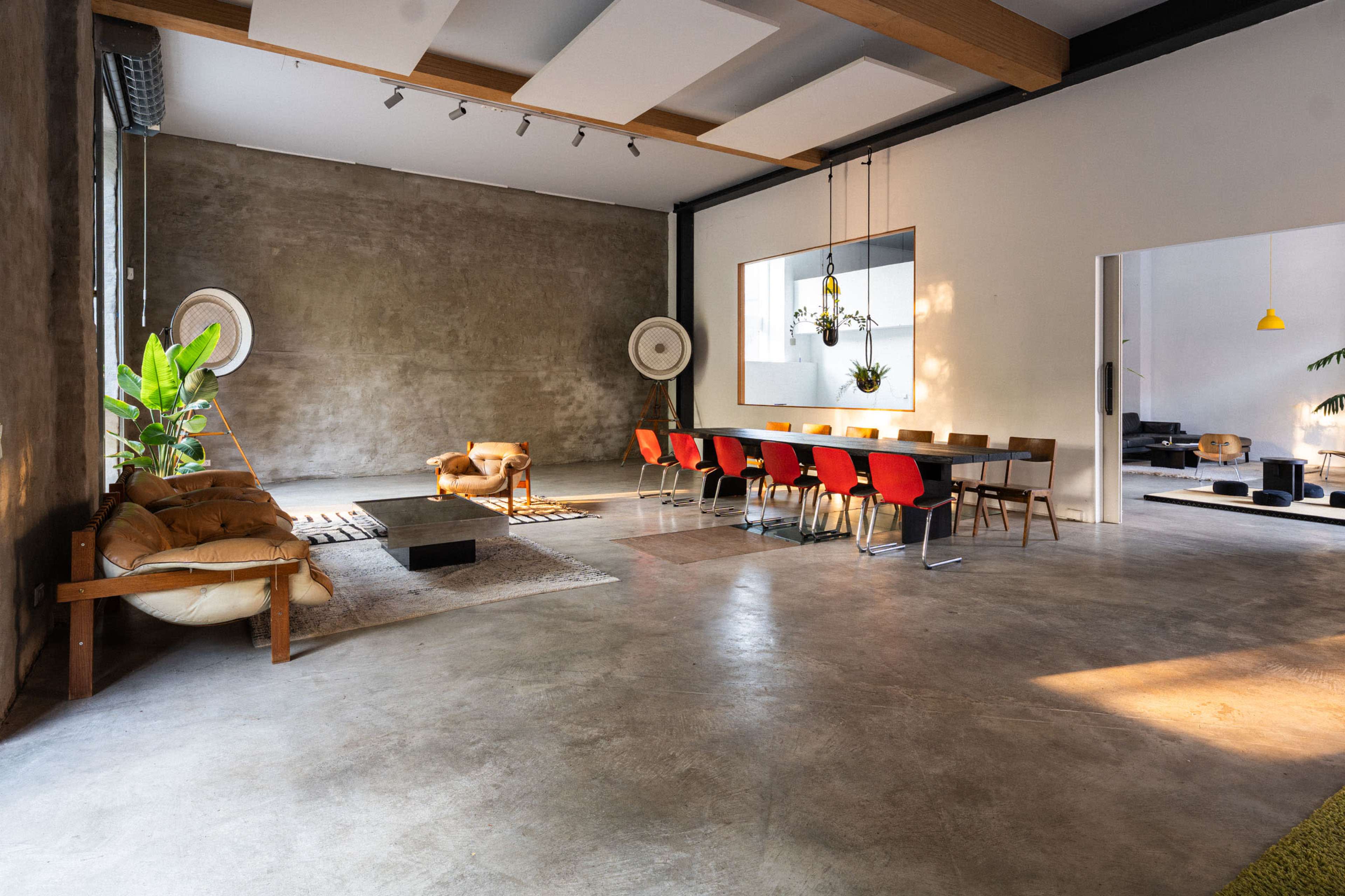 The image shows a spacious, modern interior featuring a large dining table with red chairs, a beige sofa, and green plants against a concrete wall.