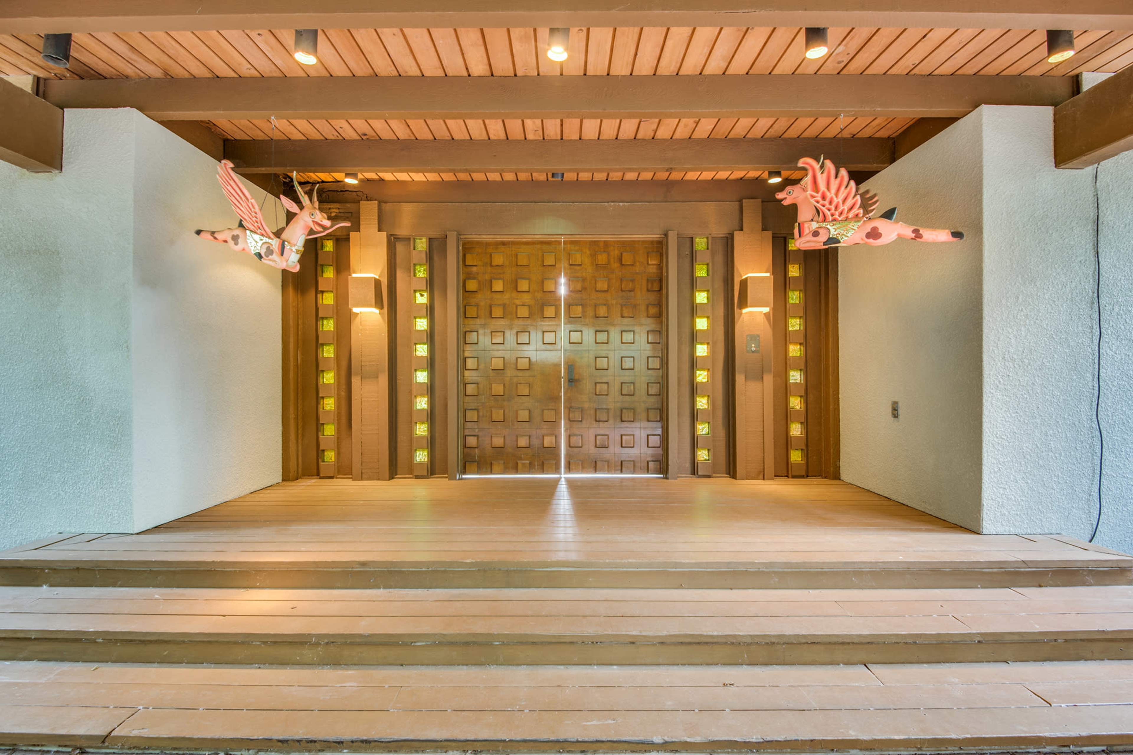A wooden entryway with a large, decorated door and two ornamental figures on either side, set against textured walls and a wooden ceiling.