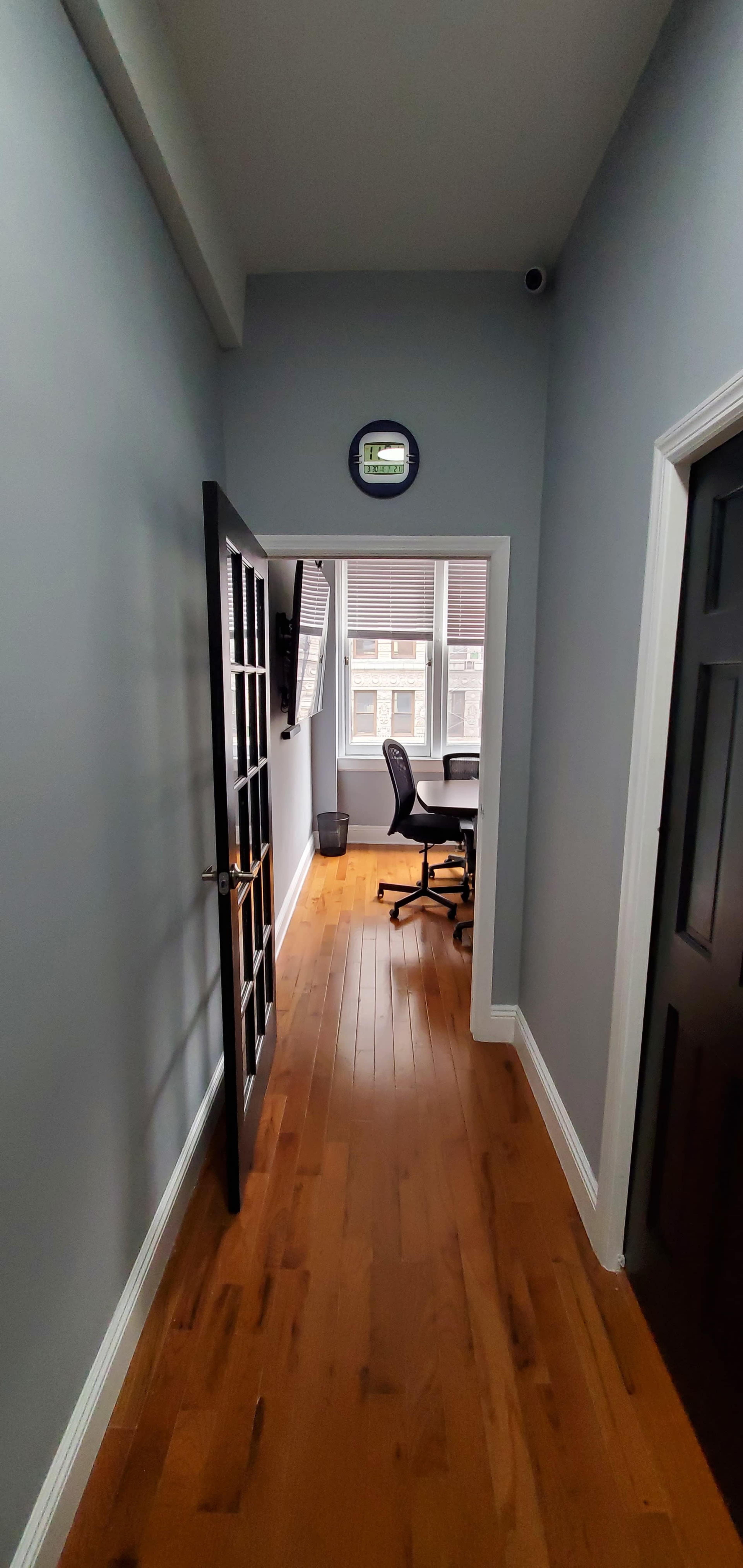 A narrow hallway leads to an office space with a desk and a chair, illuminated by natural light from a window.