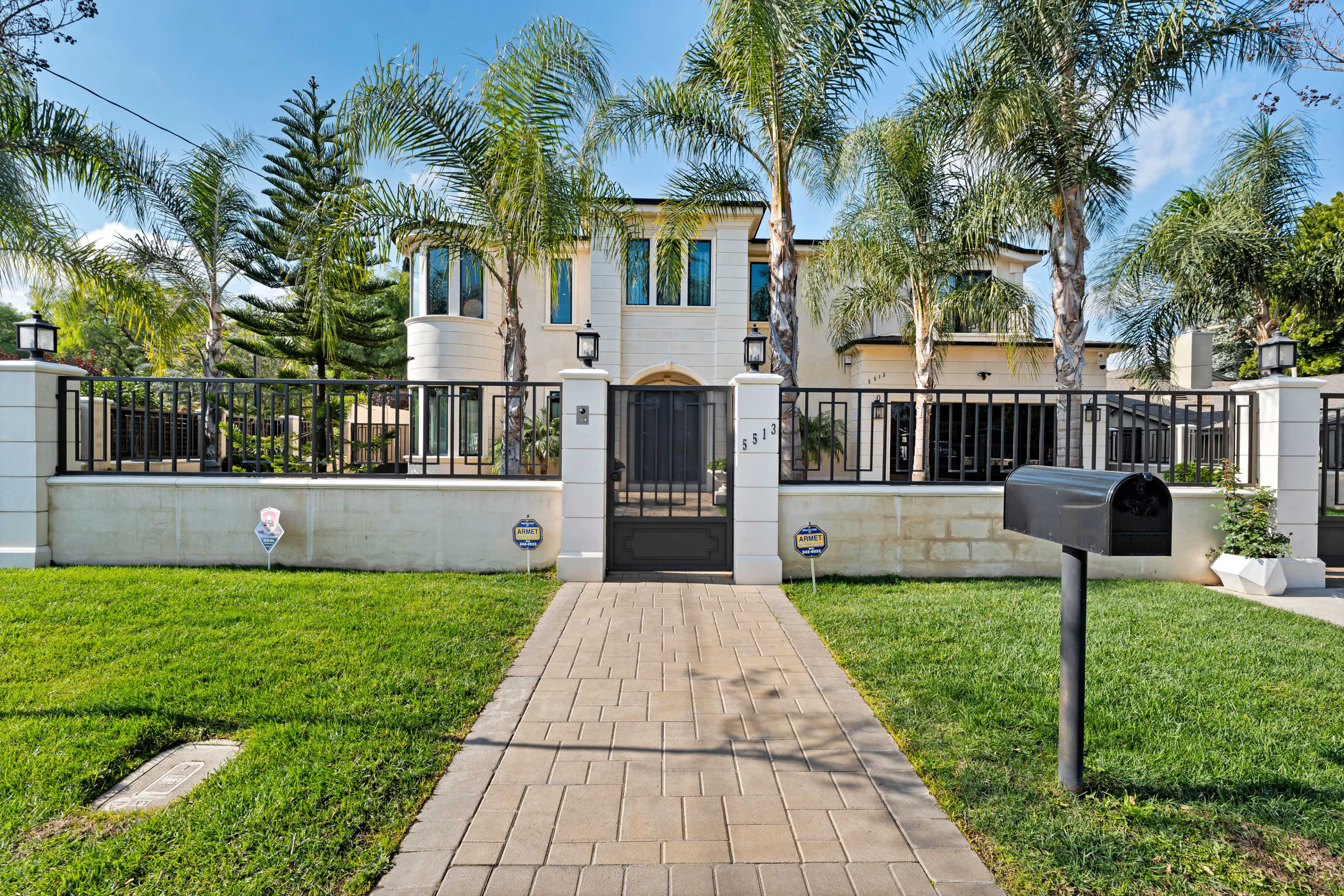 A modern two-story house with large windows is surrounded by a black metal fence and palm trees, with a paved walkway leading to the front gate.