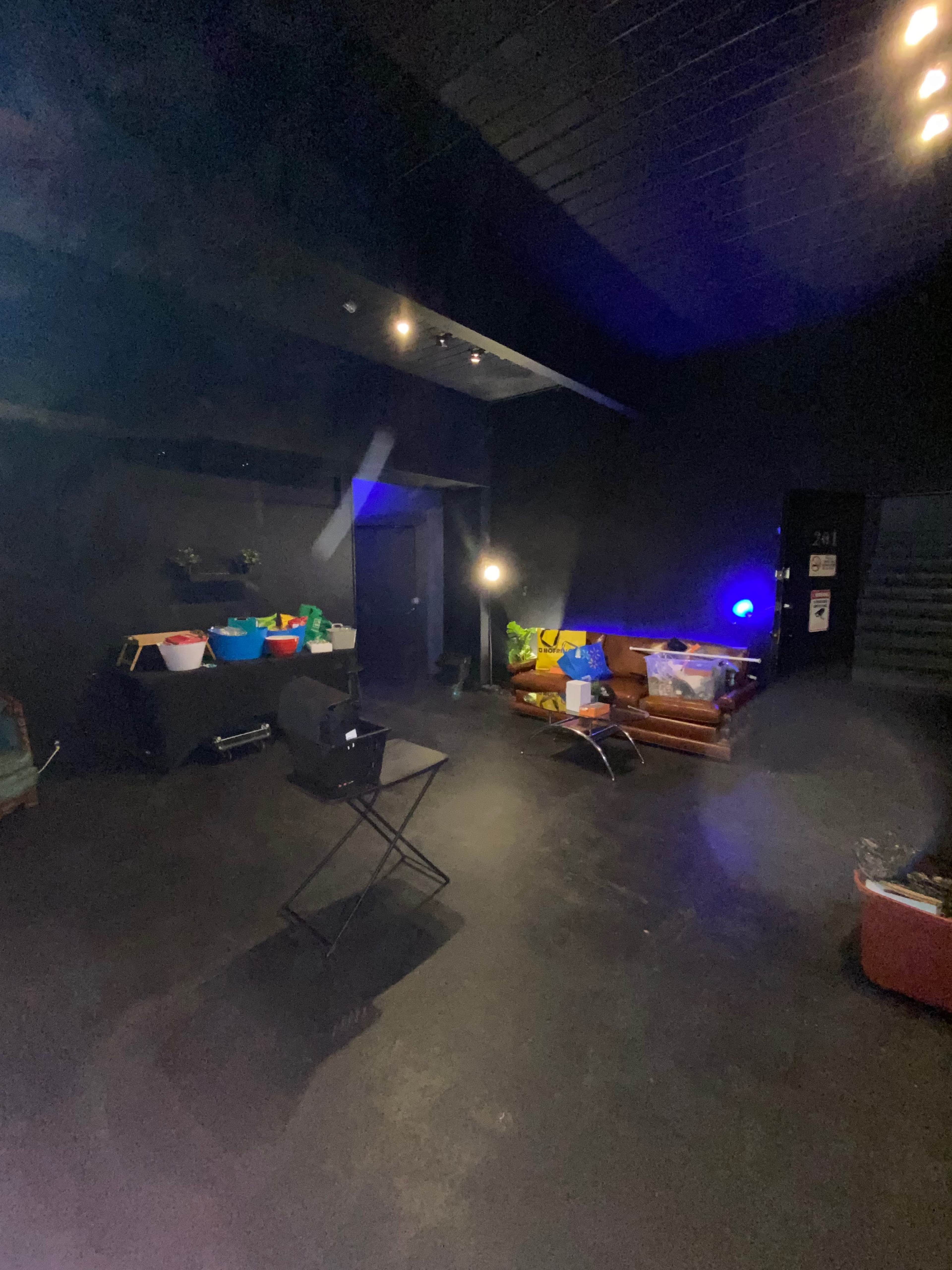 A dimly lit room with a dark floor, a chair, and various containers scattered around, along with a few light sources illuminating different areas.
