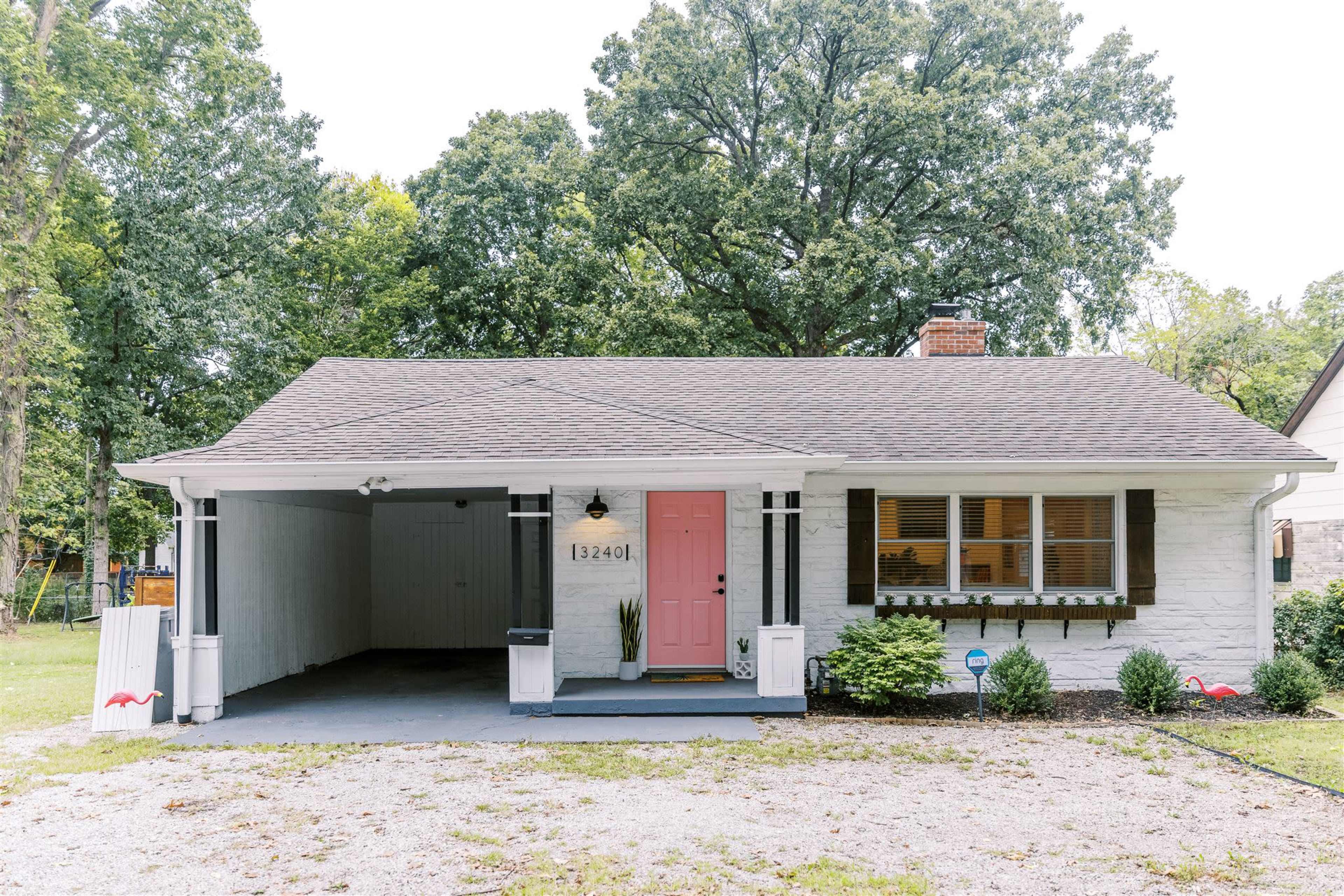 A single-story house with a pink front door and a covered carport, surrounded by greenery and gravel landscaping.
