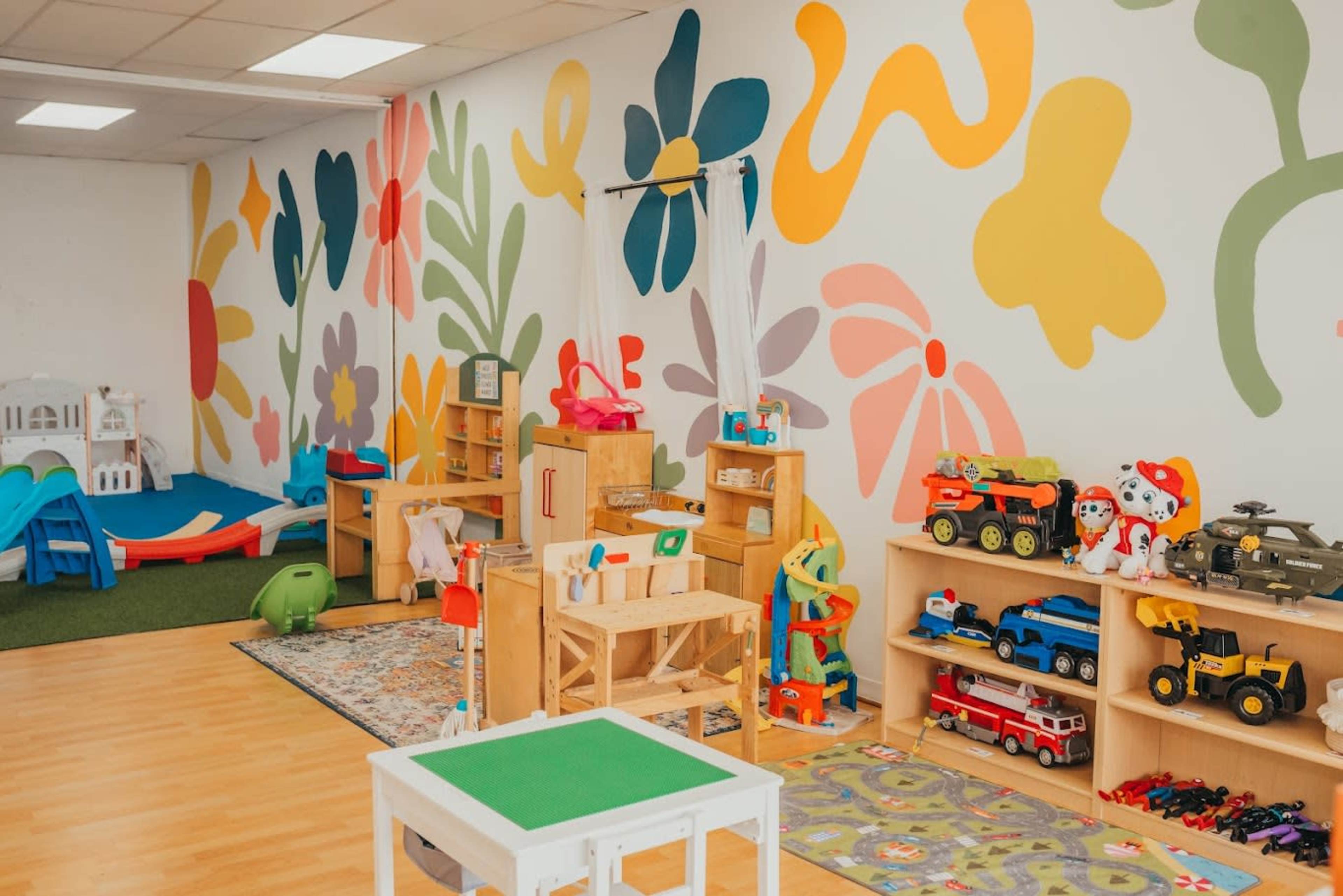The image shows a brightly colored playroom with a variety of toys, furniture, and a mural of flowers on the wall.