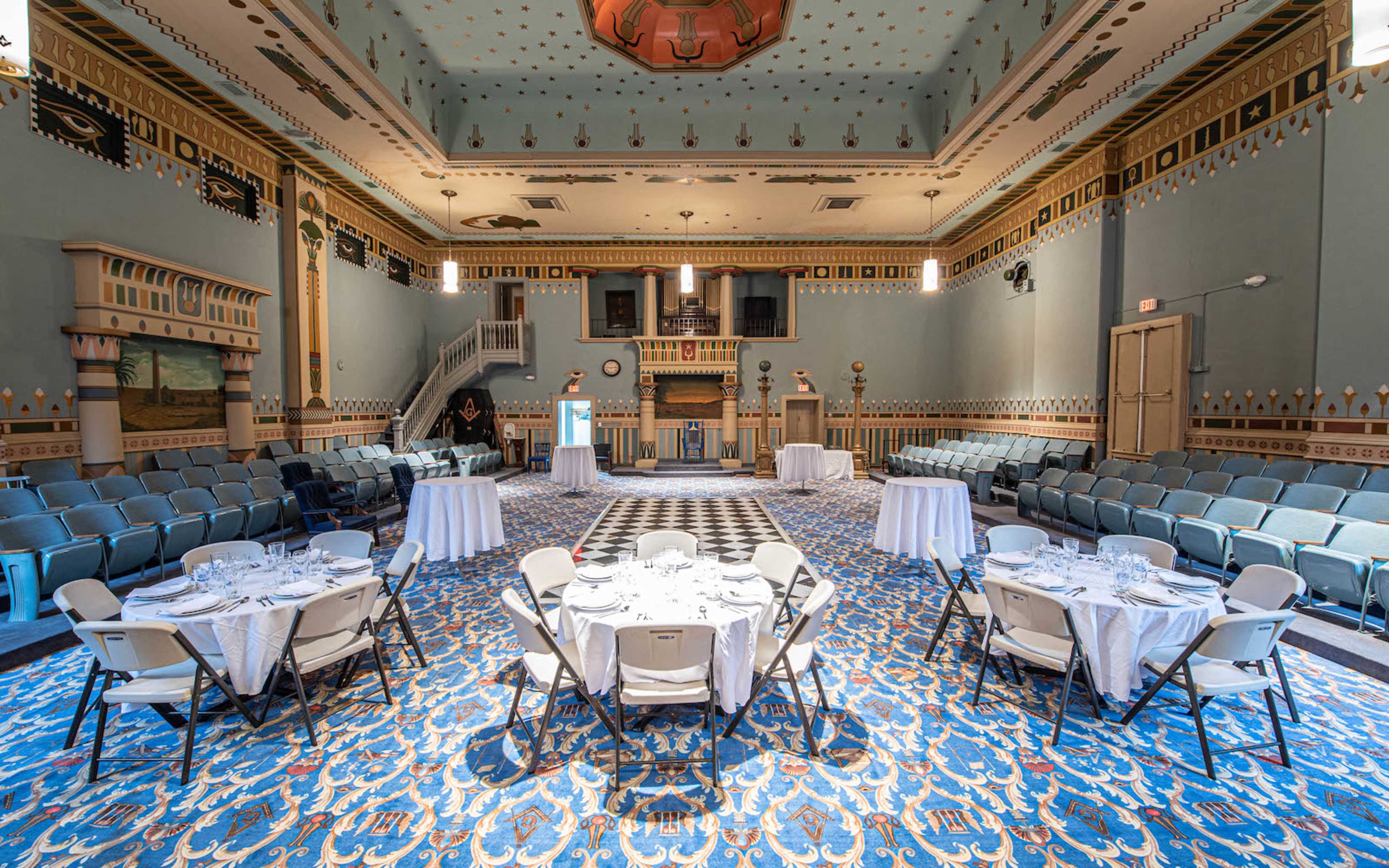 A spacious, elegantly decorated hall with circular tables set for dining, surrounded by rows of chairs and ornate walls.
