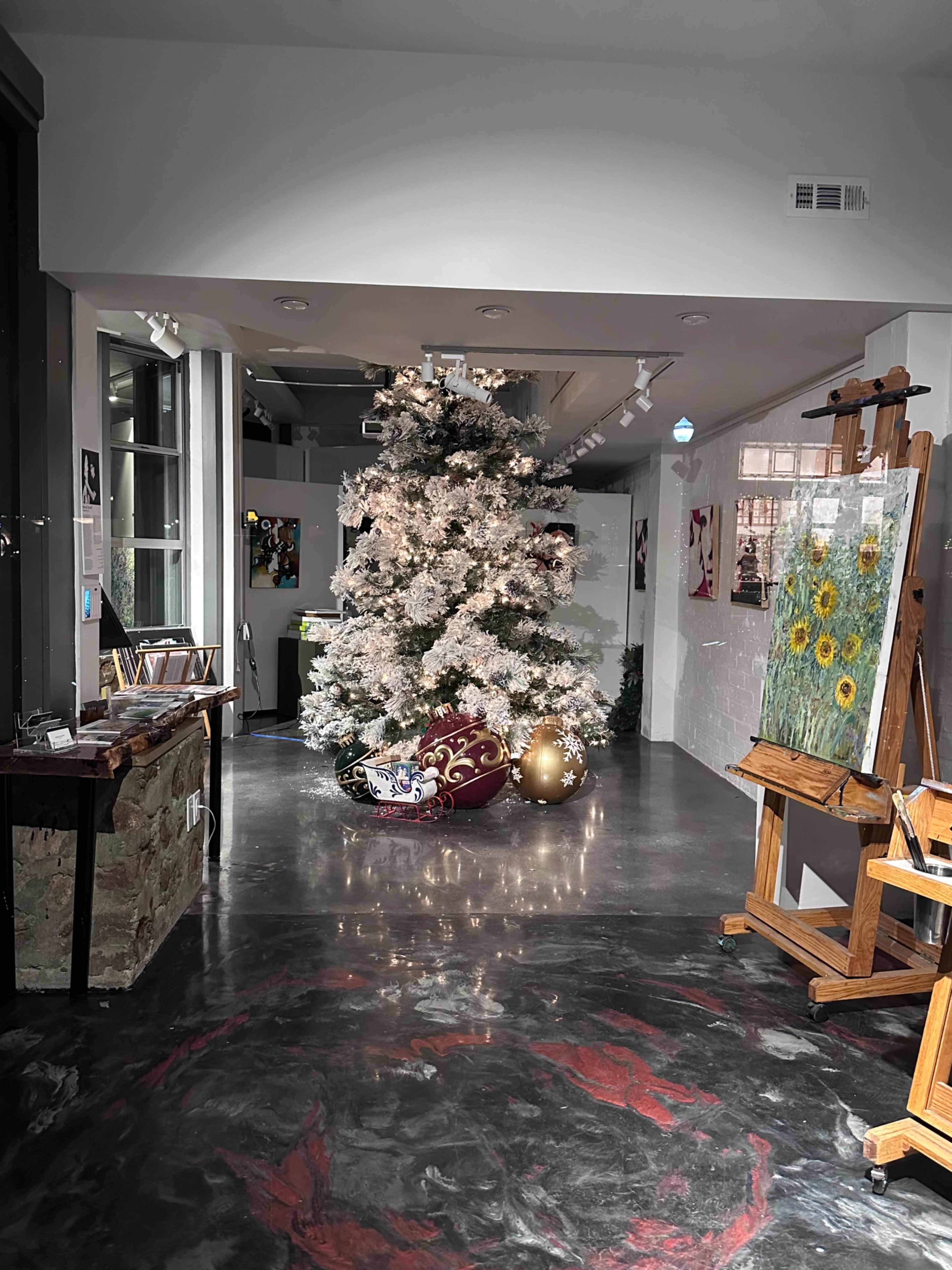 A white Christmas tree is decorated with lights and ornaments in an art gallery, surrounded by various artworks and an easel.