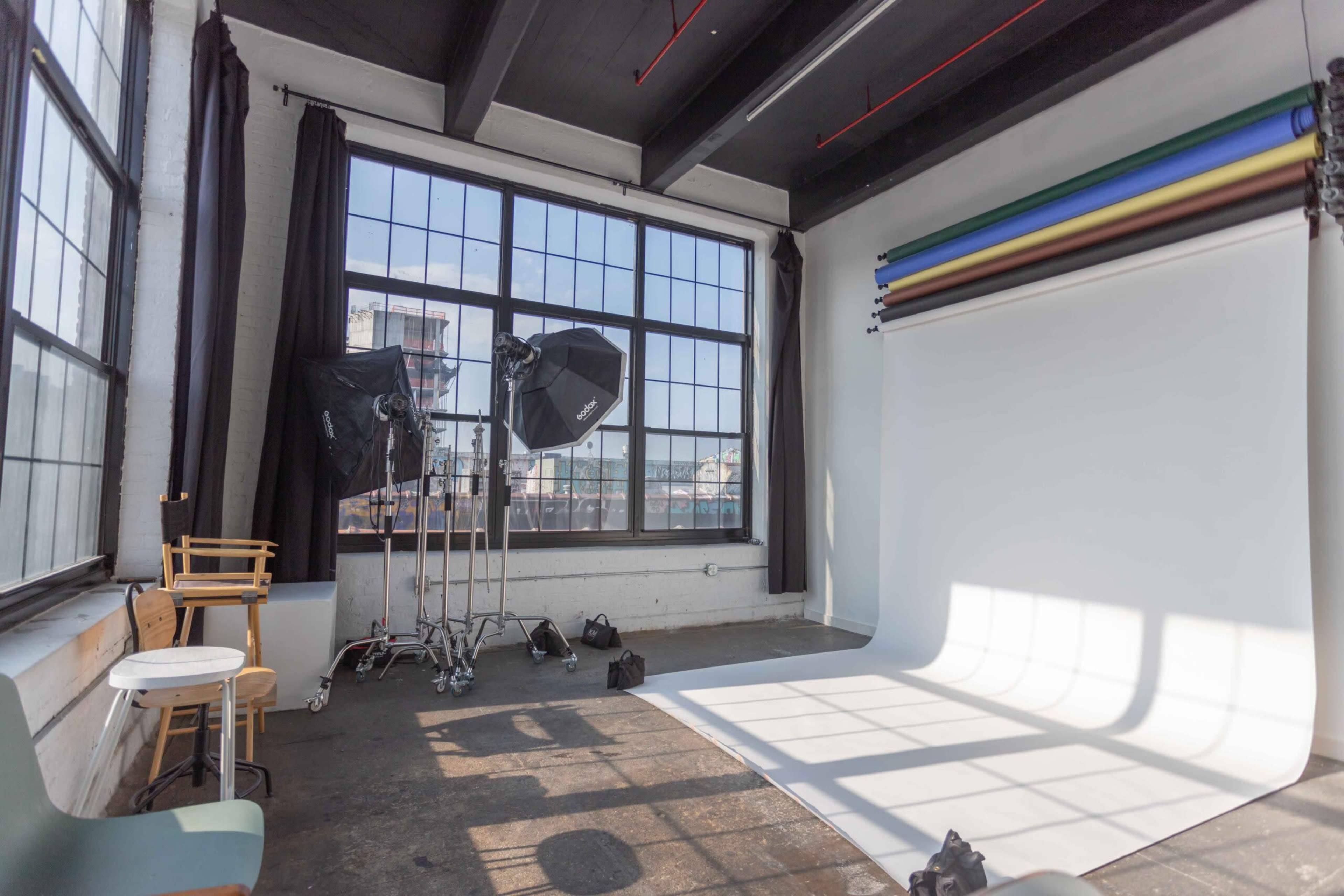 The image shows a photography studio with large windows, backdrop rolls, lighting equipment, and an empty area for capturing images.