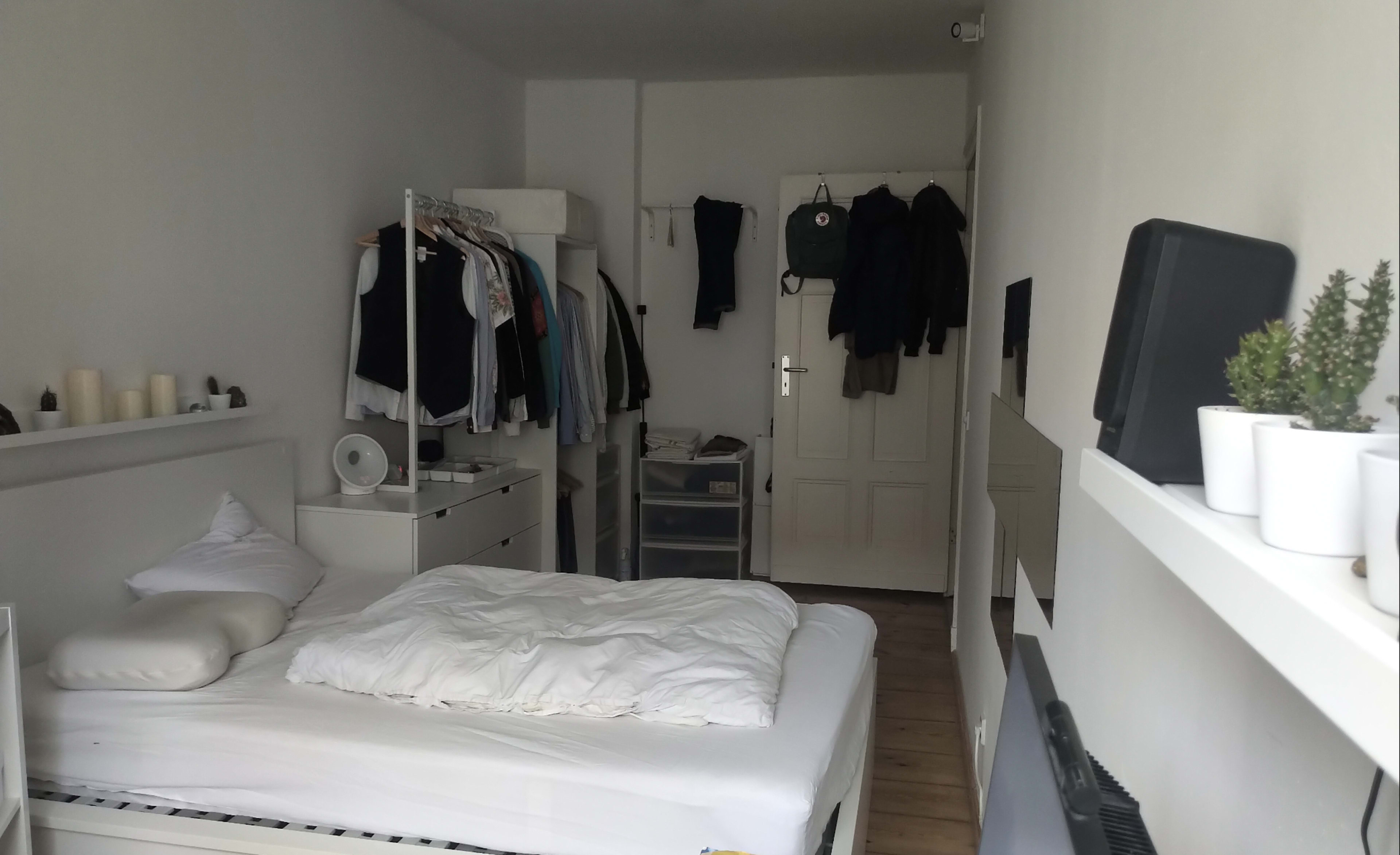 The room features a bed with white bedding, a clothing rack with hanging garments, a dresser, and a door leading to another area, with clothing hung on the back of it.