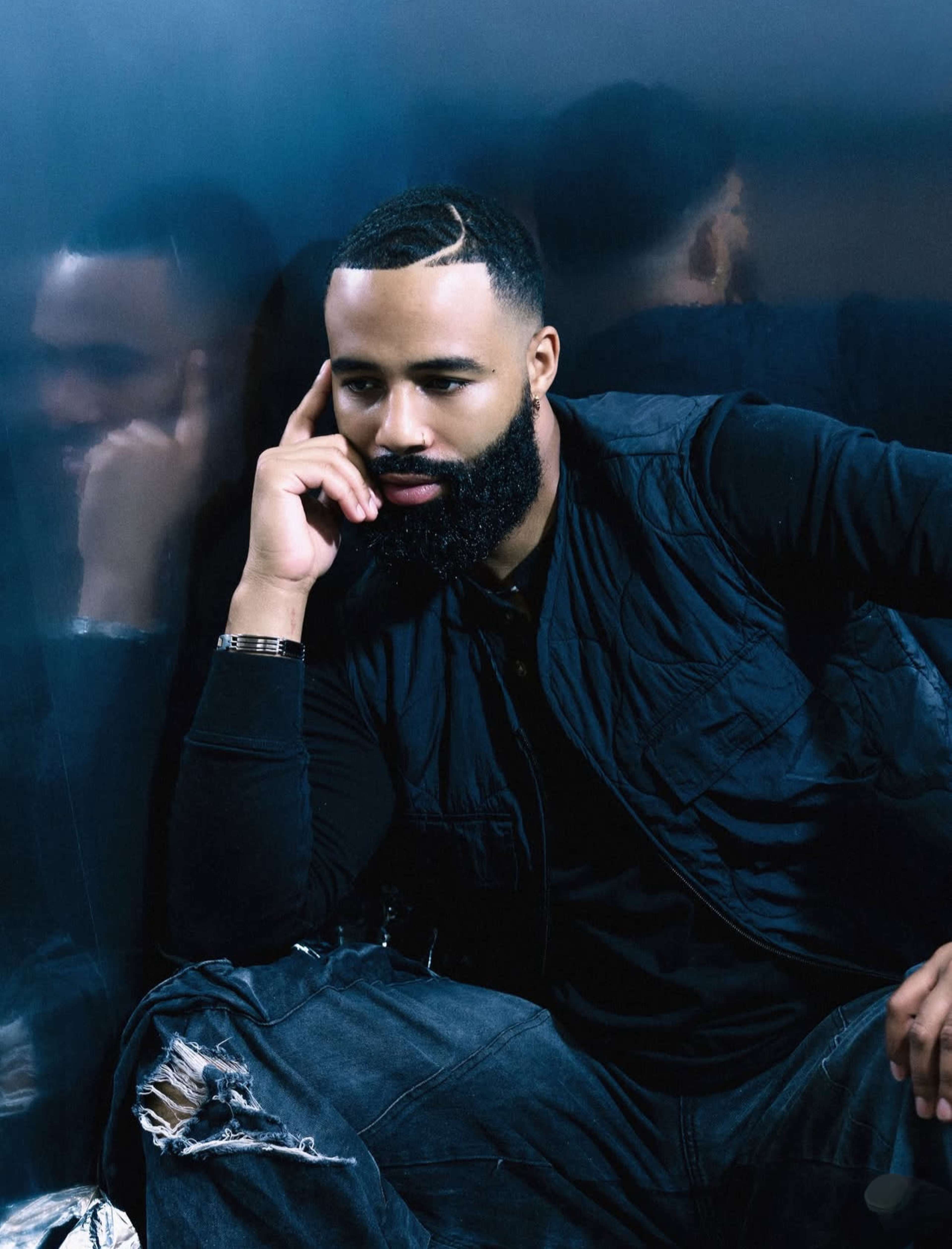 A man with a beard is sitting against a reflective surface, wearing a black vest and distressed jeans.