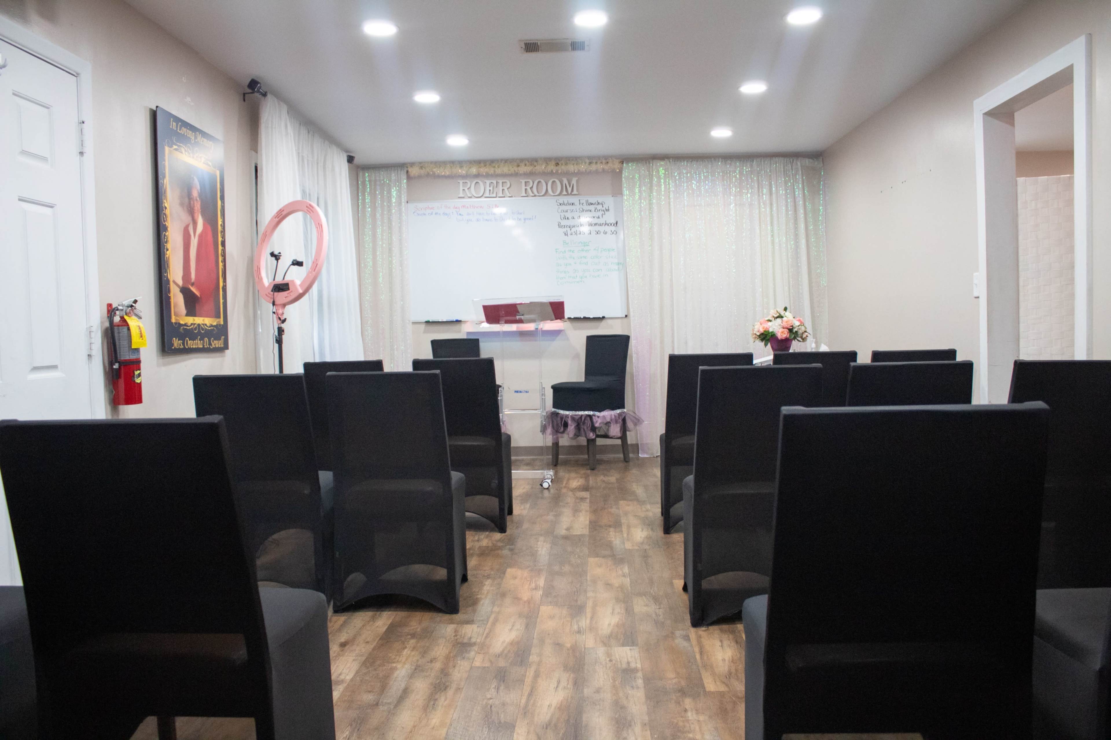 The image shows a small, well-lit room arranged with black chairs neatly placed in rows facing a whiteboard and a small table at the front.