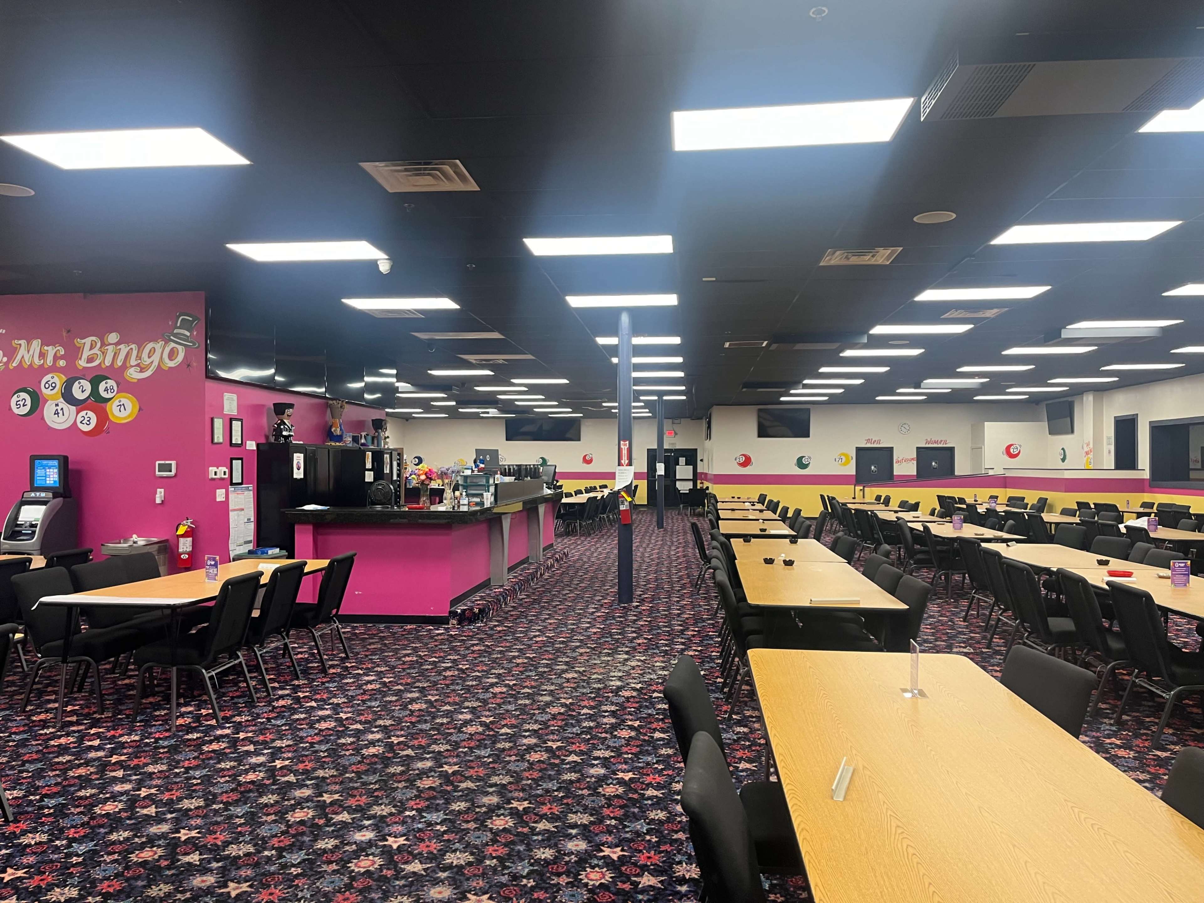 A spacious bingo hall with several rows of tables and chairs, a bar area, and vibrant purple walls decorated with casino-themed elements.