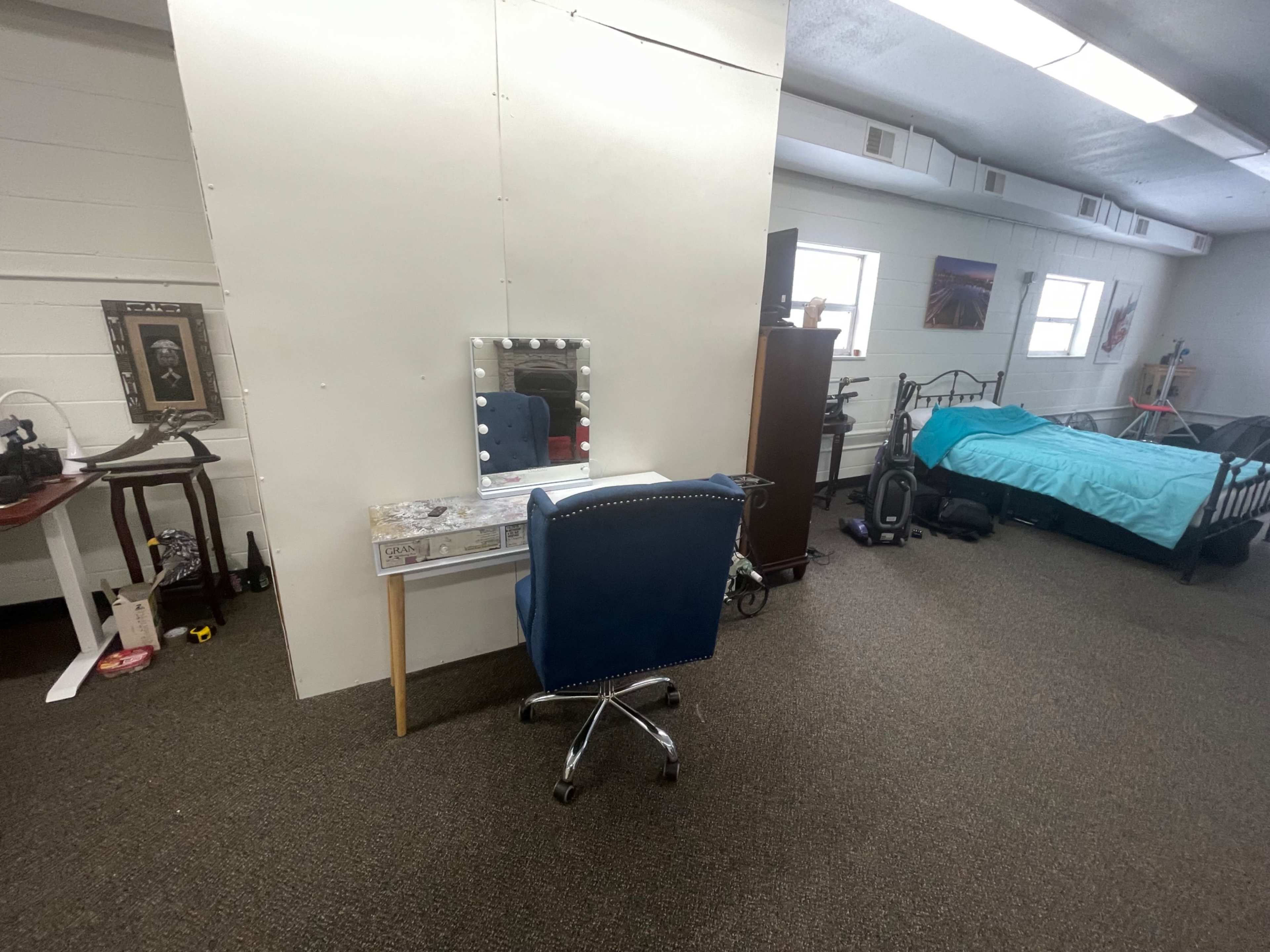 A simple room featuring a bed, a vanity with a mirror and chair, a dresser, and various items on the floor.