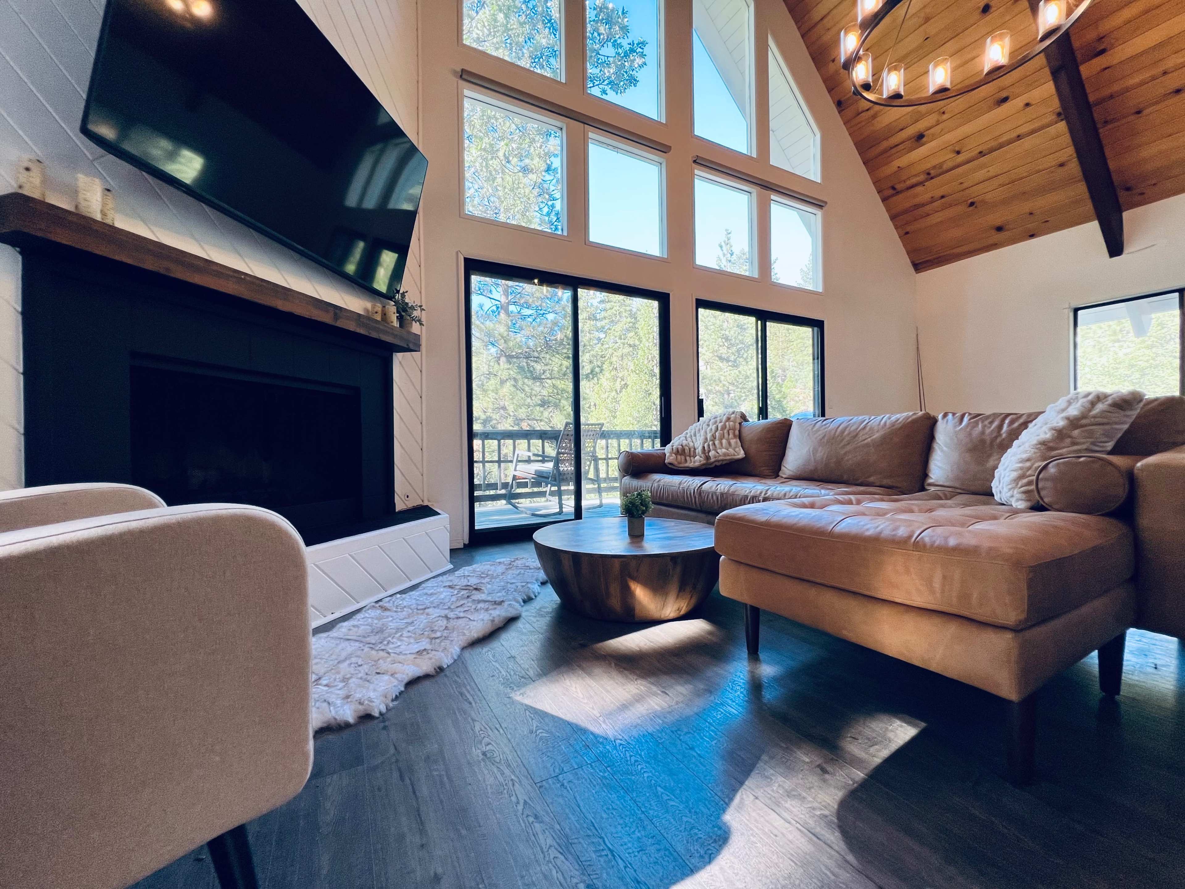 The image shows a cozy living room with a large window, a sectional sofa, a circular coffee table, and a fireplace, all illuminated by natural light.