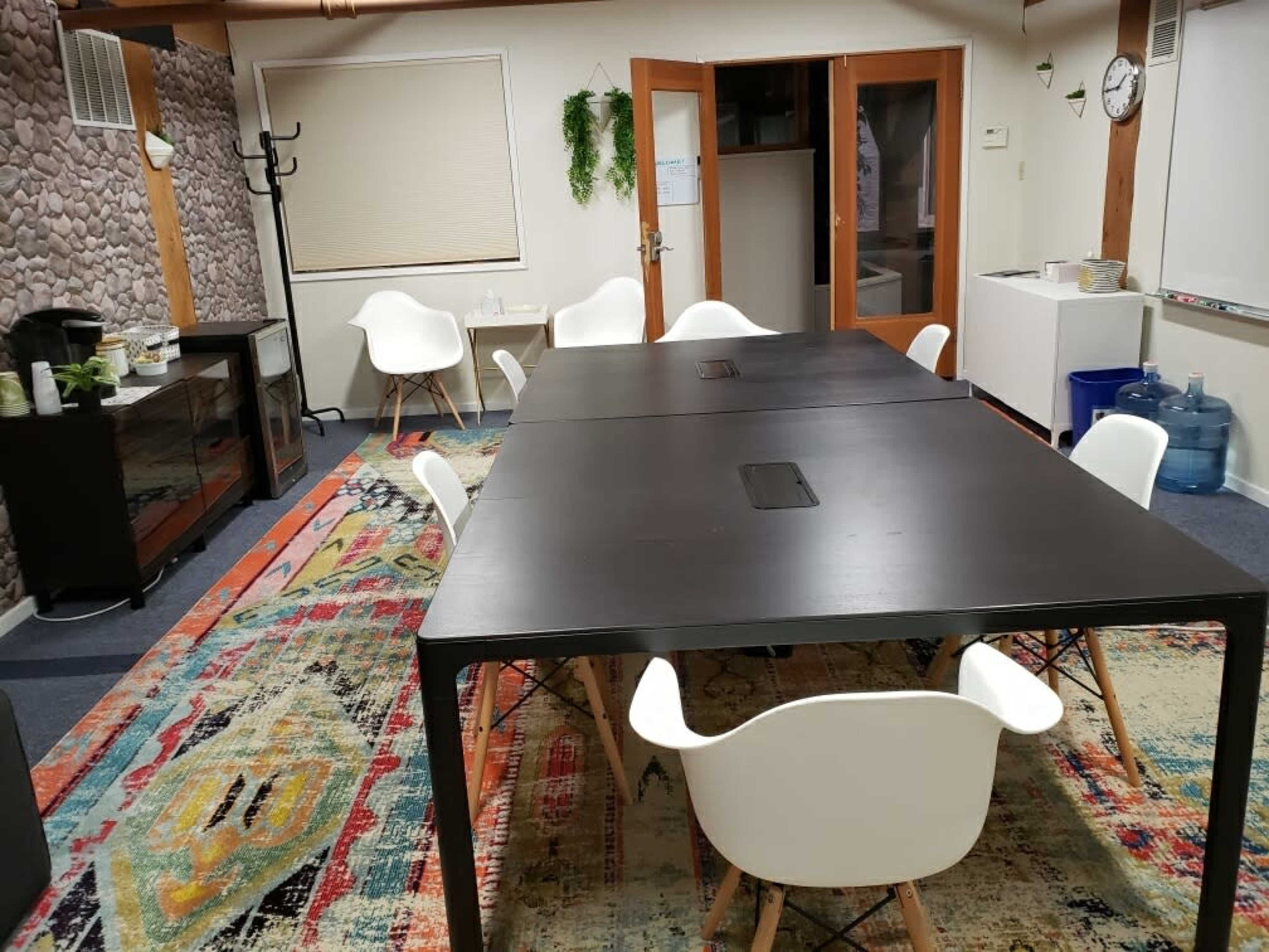 A modern office meeting room with two large black tables, white chairs, and a colorful patterned rug.