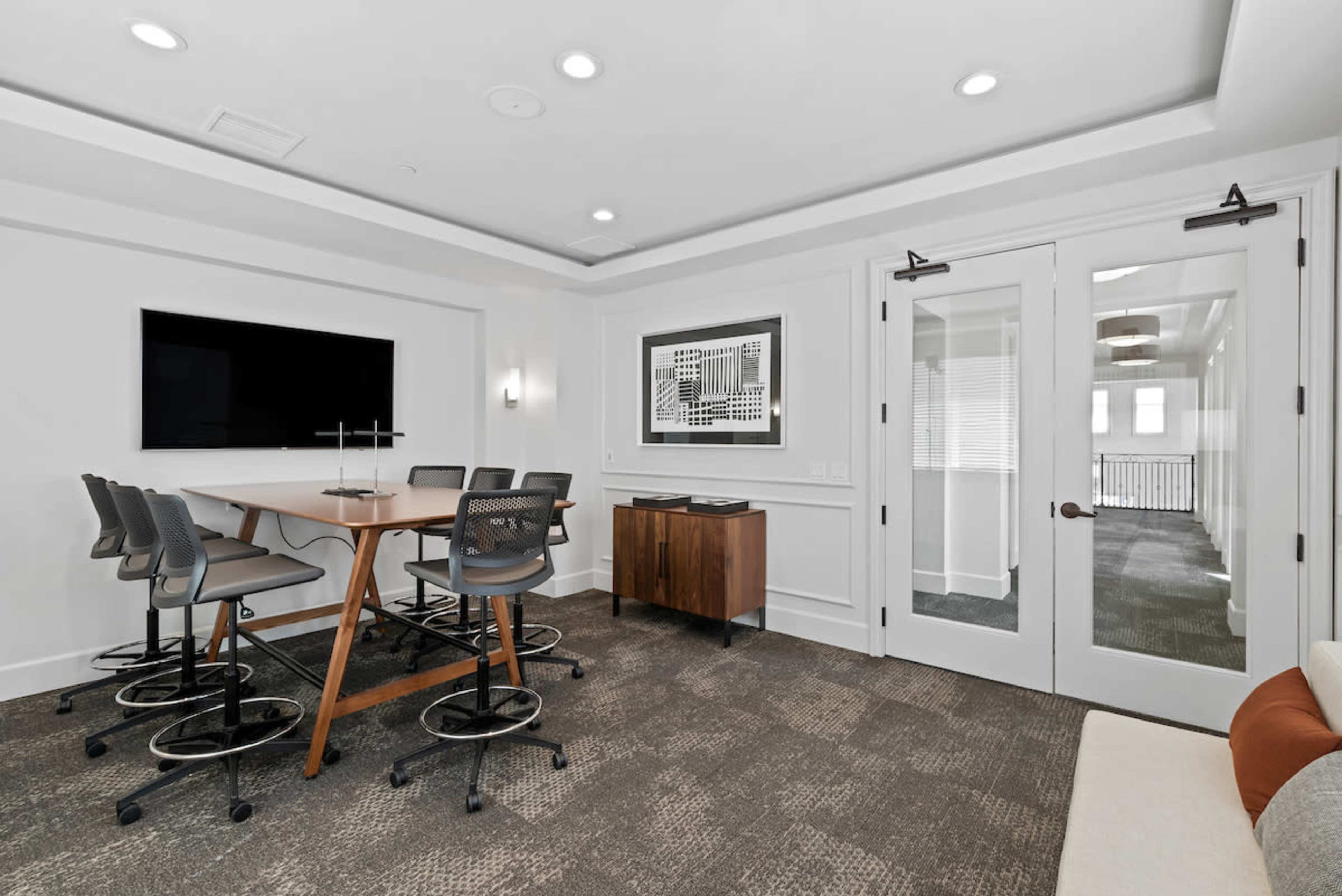 A modern conference room features a large table surrounded by high-backed chairs, a TV mounted on the wall, and glass doors leading to another area.