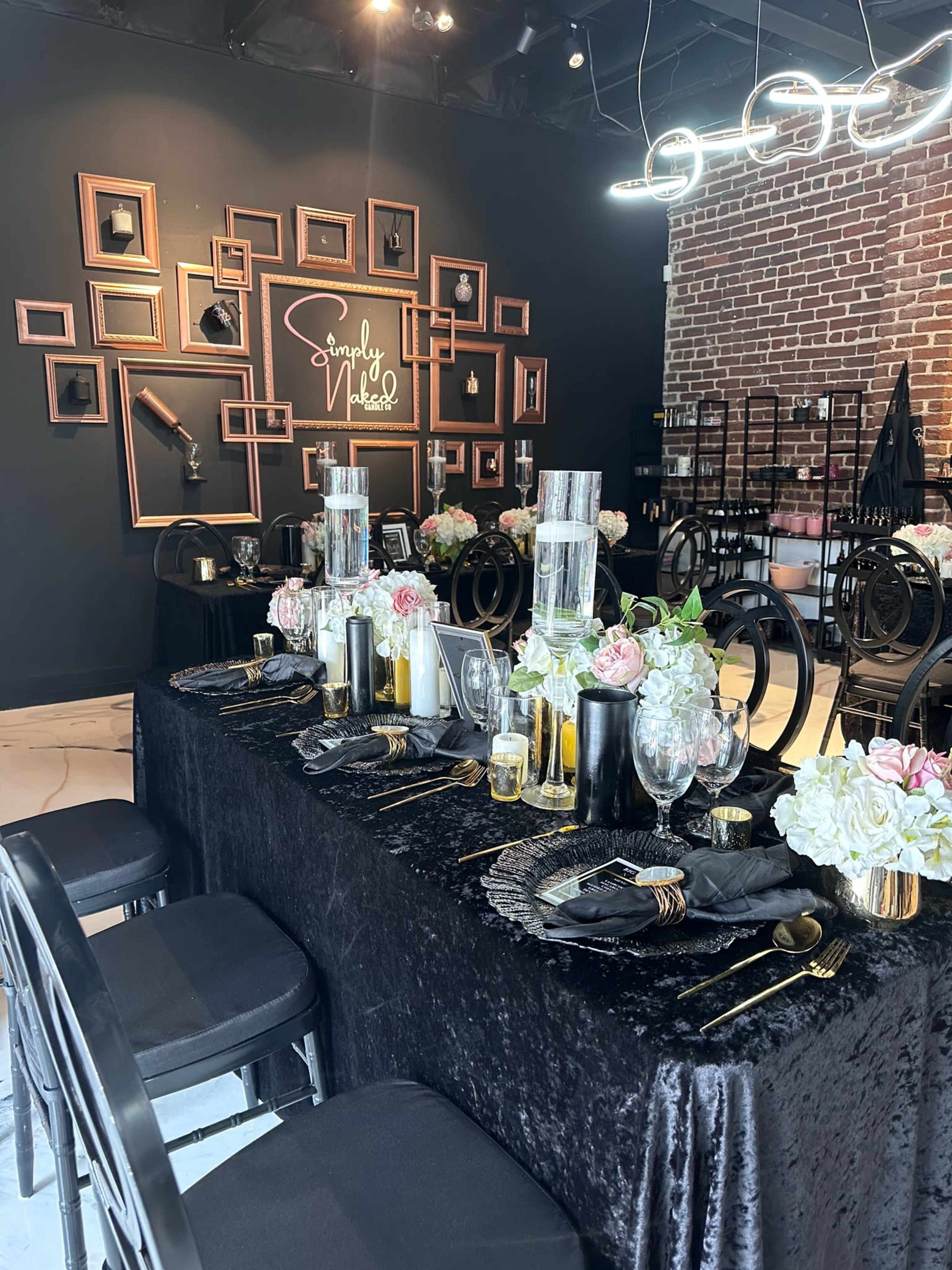 A dining table elegantly set with black and gold tableware, surrounded by black chairs and adorned with floral centerpieces against a dark wall featuring framed pictures.