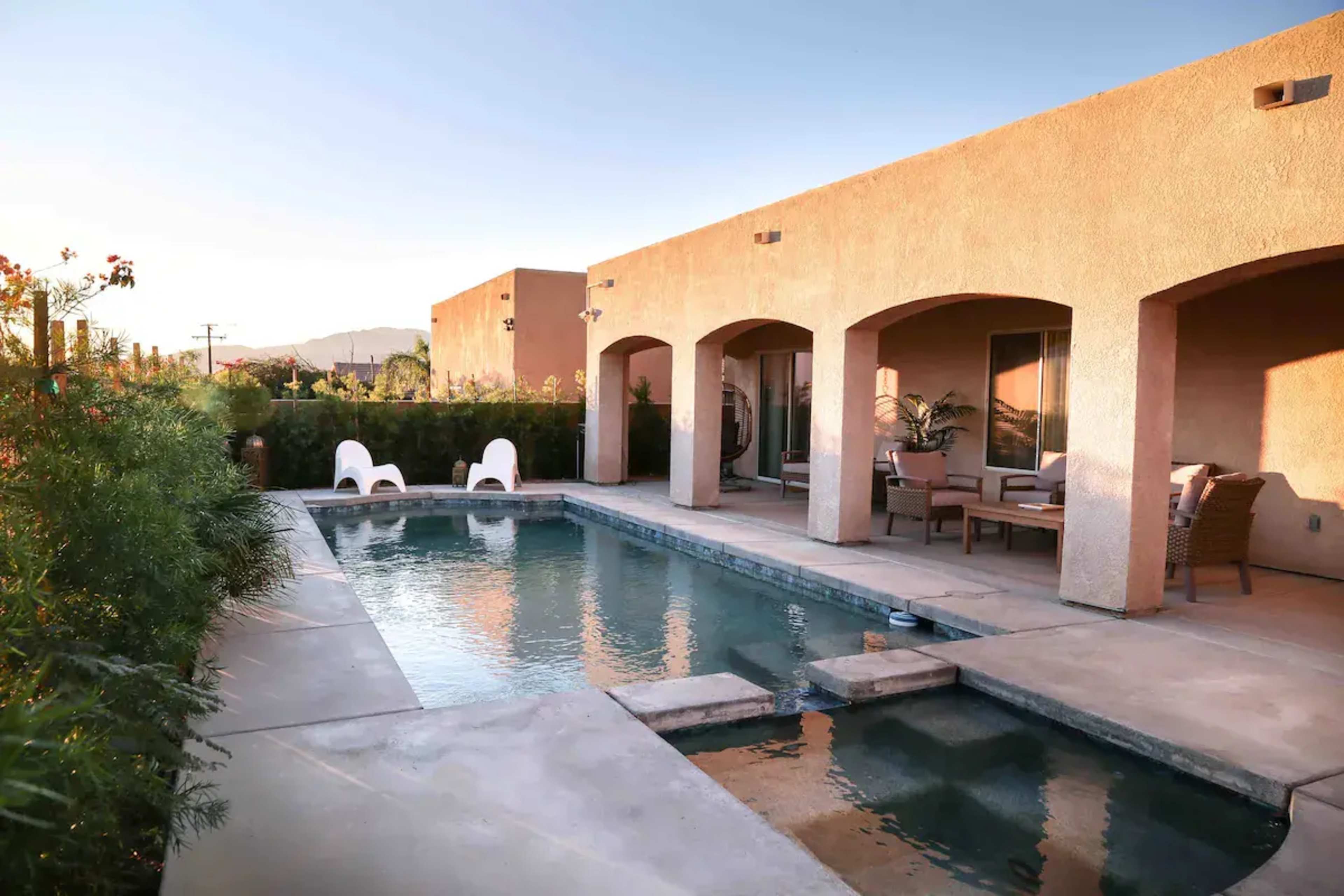 The image shows a swim pool surrounded by a patio with lounge chairs and a sofa, adjacent to a modern stucco building.