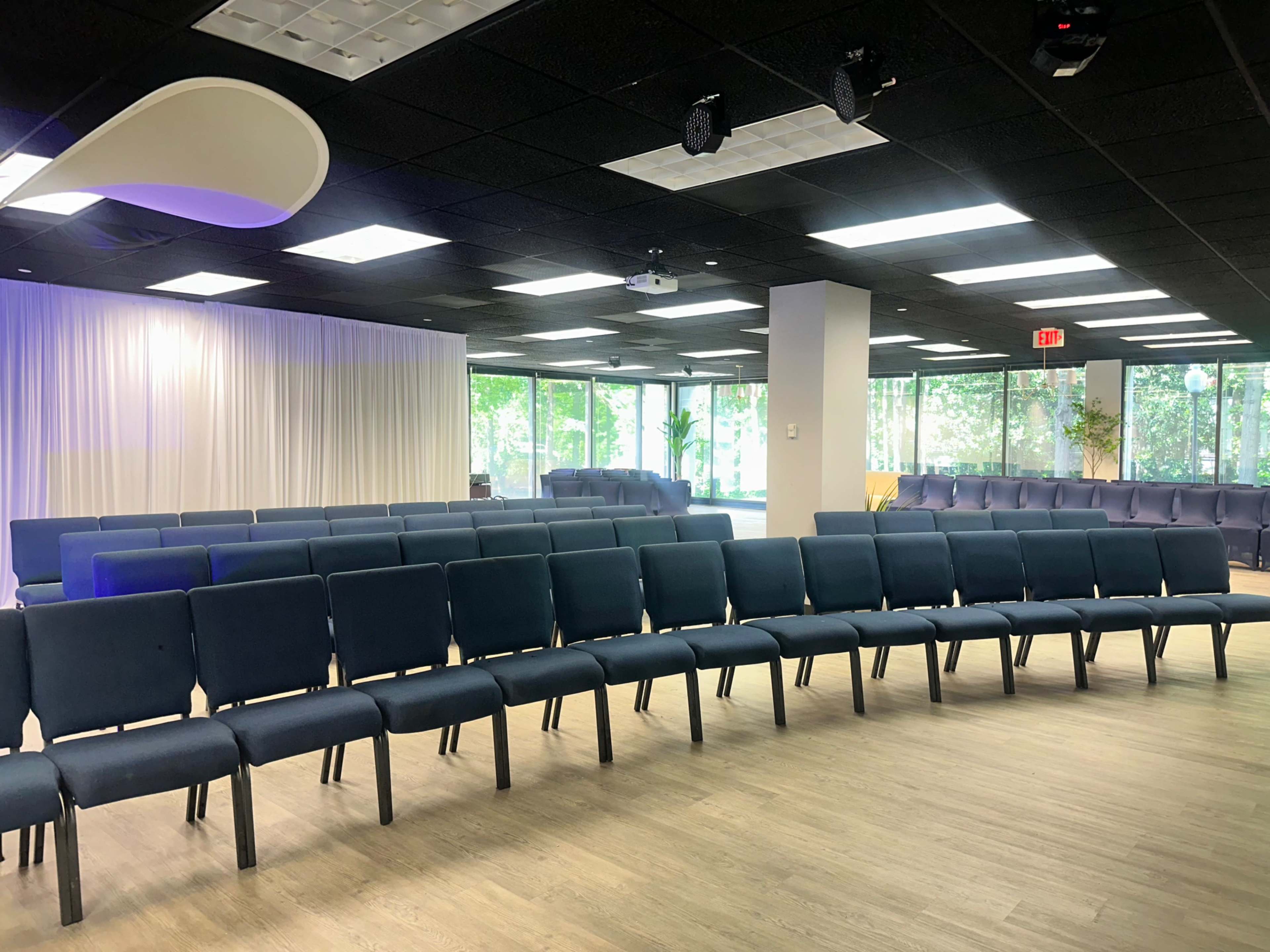The image shows a spacious room set up with rows of chairs facing a stage, with large windows allowing natural light to enter.