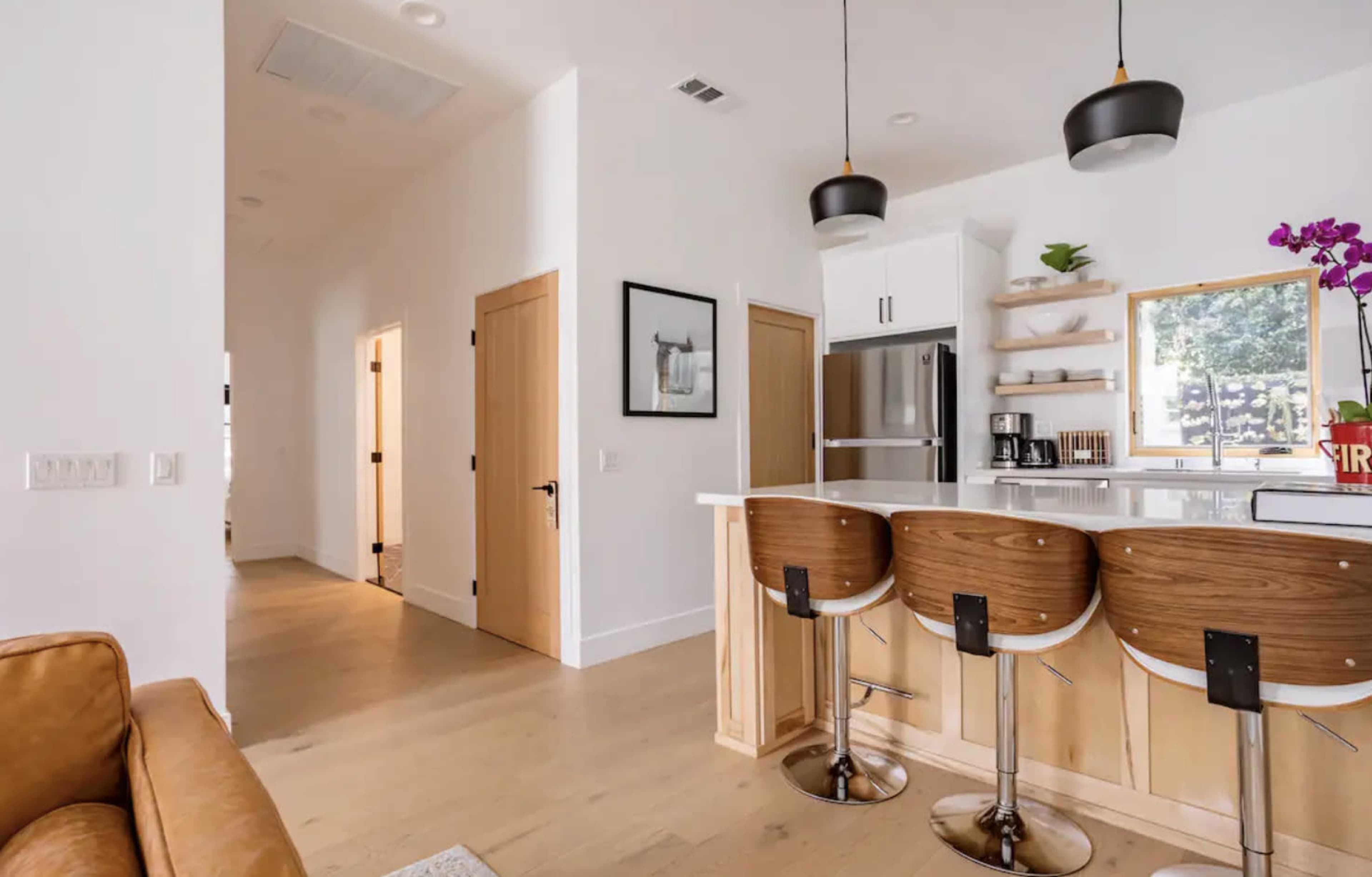 A modern kitchen with two barstools, a refrigerator, and a corridor leading to other rooms.