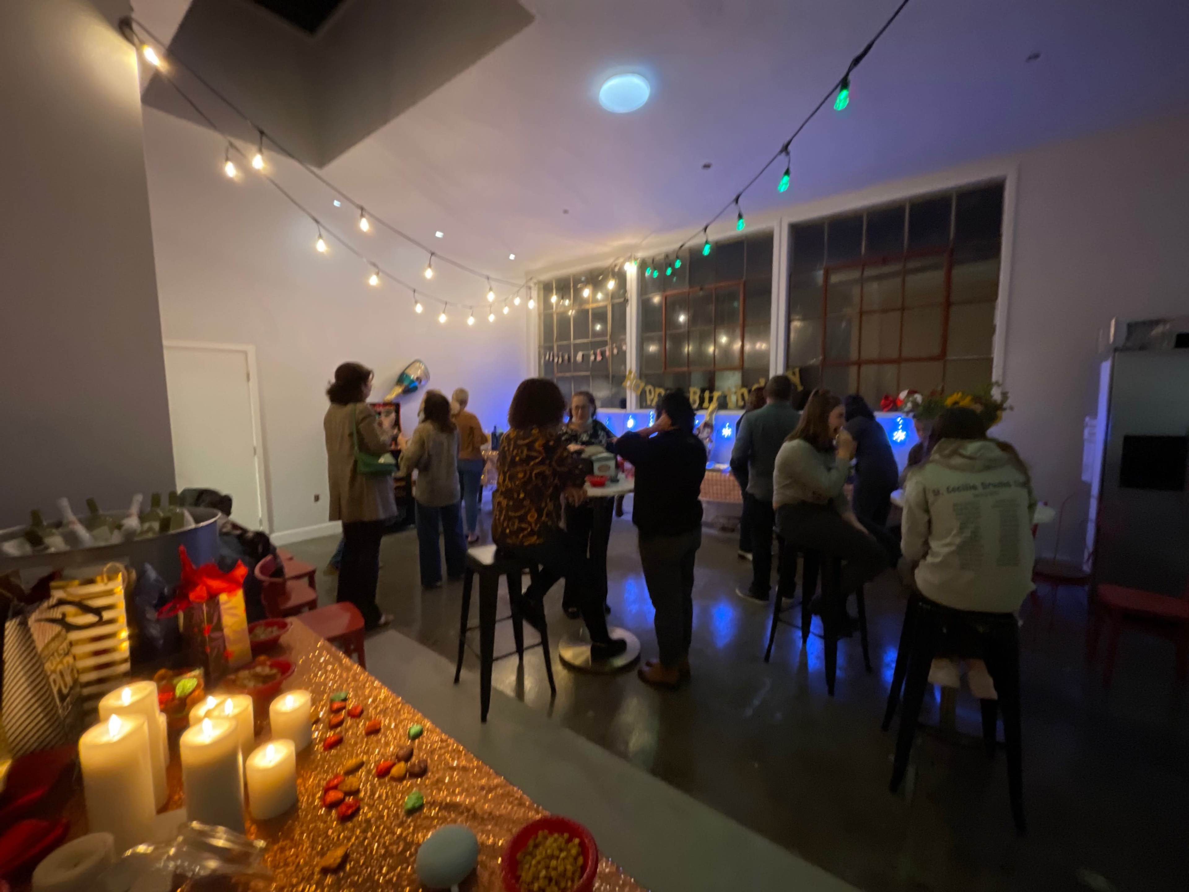 A group of people is gathered in a warmly lit venue with a bar, surrounded by festive decorations and glowing string lights.