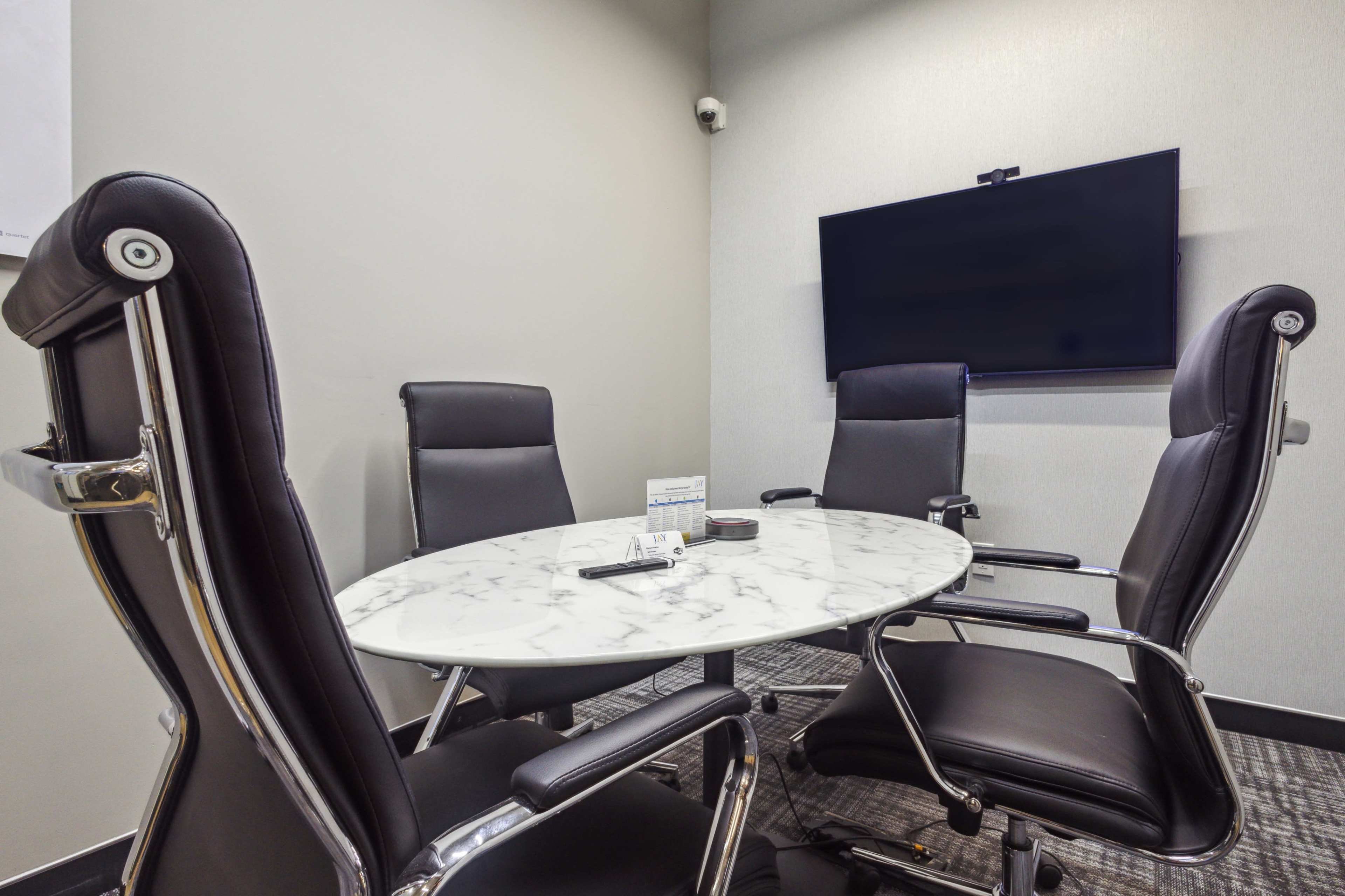 A small, modern conference room features a round marble table surrounded by four black executive chairs and a wall-mounted TV.