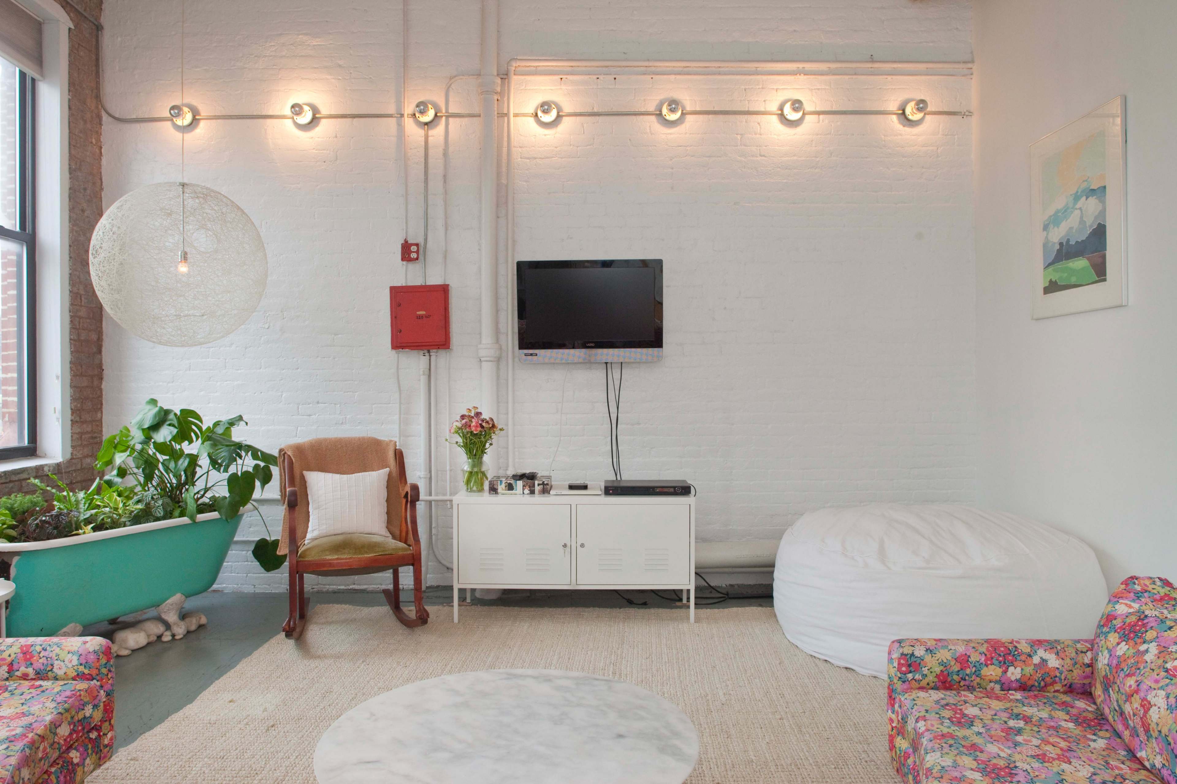 A modern living space with a white brick wall, a television mounted on the wall, a large light fixture, a green bathtub holding plants, and various seating options including a floral couch and a bean bag chair.