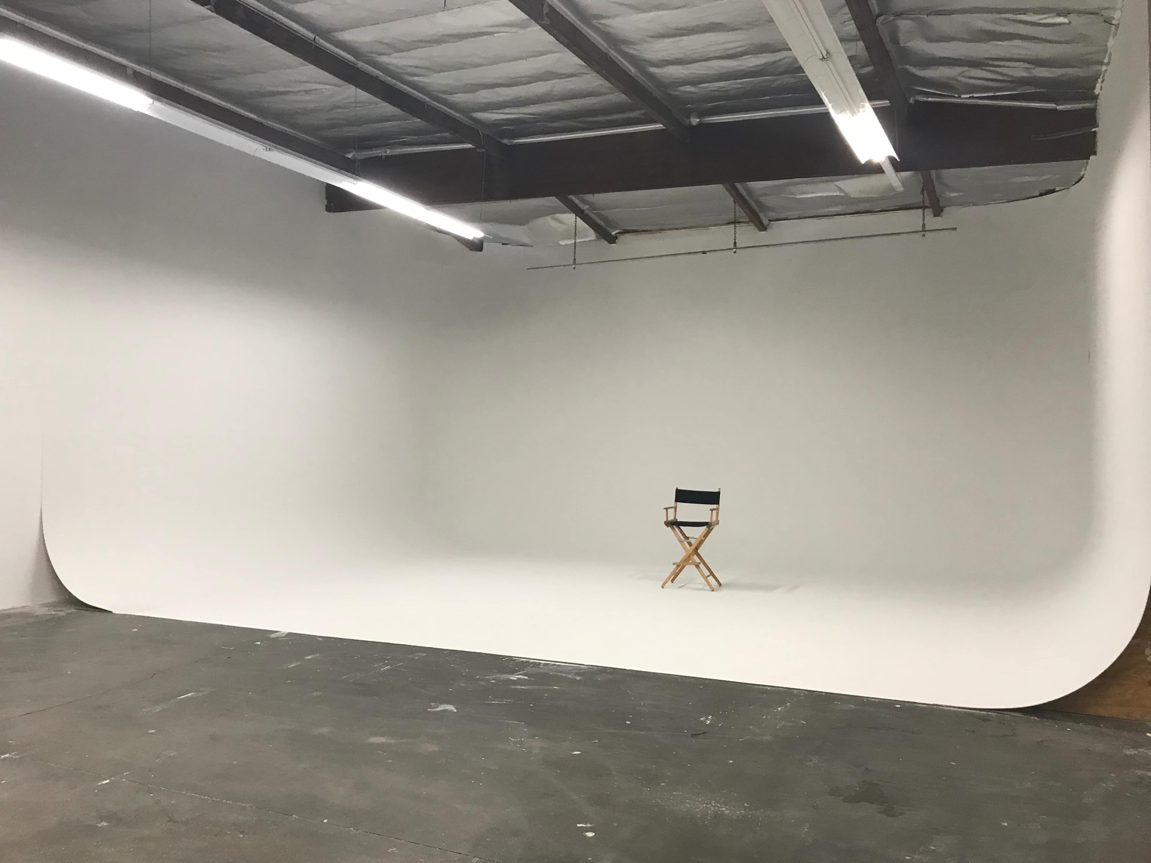 A wooden director's chair is centered in an empty white studio space with a curved backdrop.