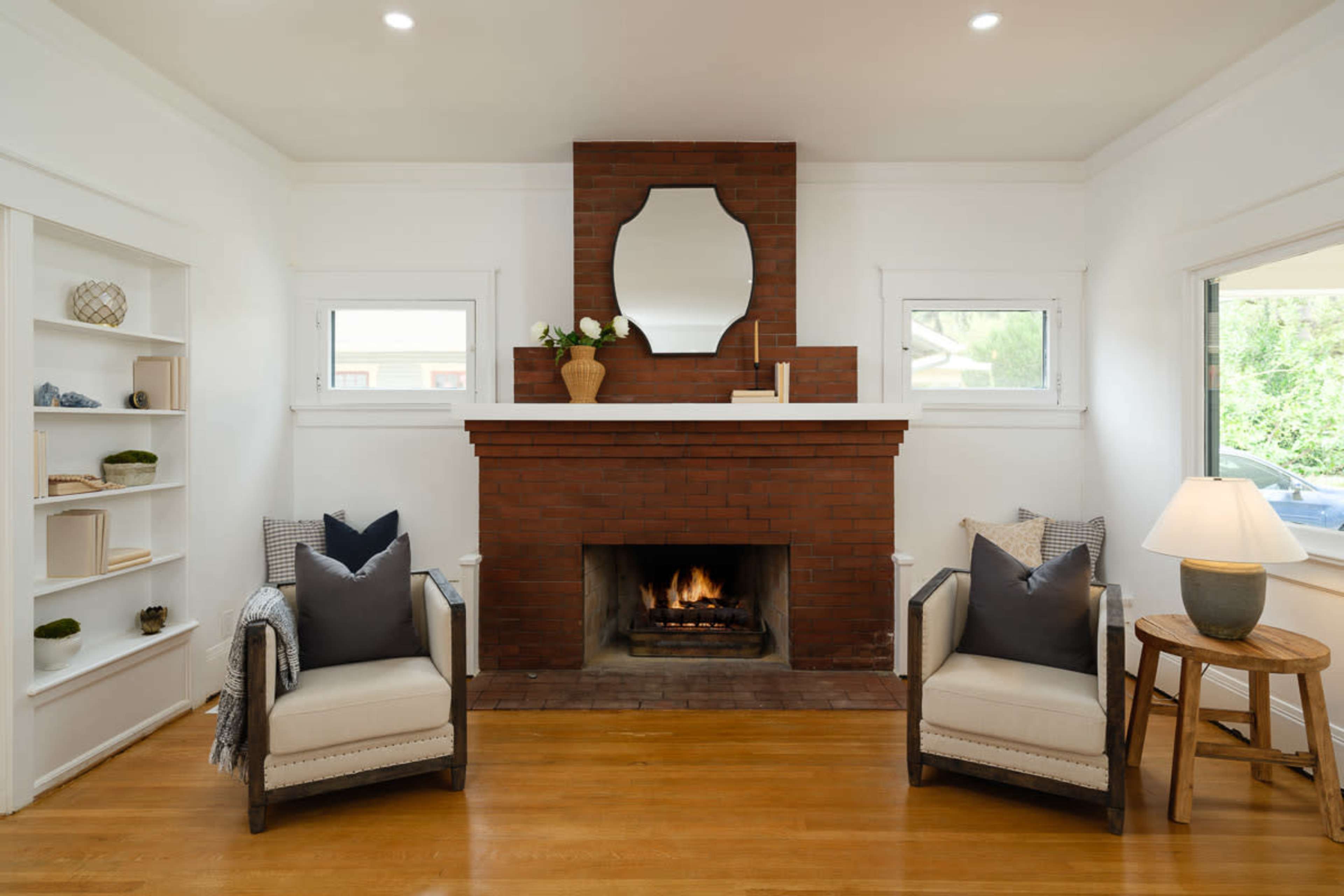 A cozy living room features a brick fireplace with a mirror above it, flanked by two armchairs and a wooden side table with a lamp.