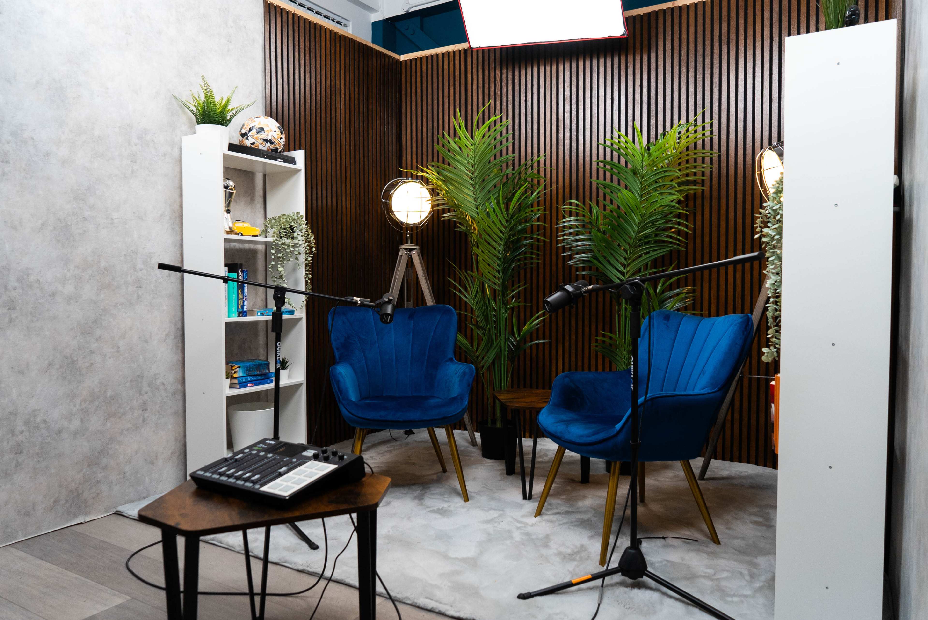 A media studio setup featuring two blue chairs, a wooden coffee table, a sound mixer, and potted plants against a wooden backdrop.