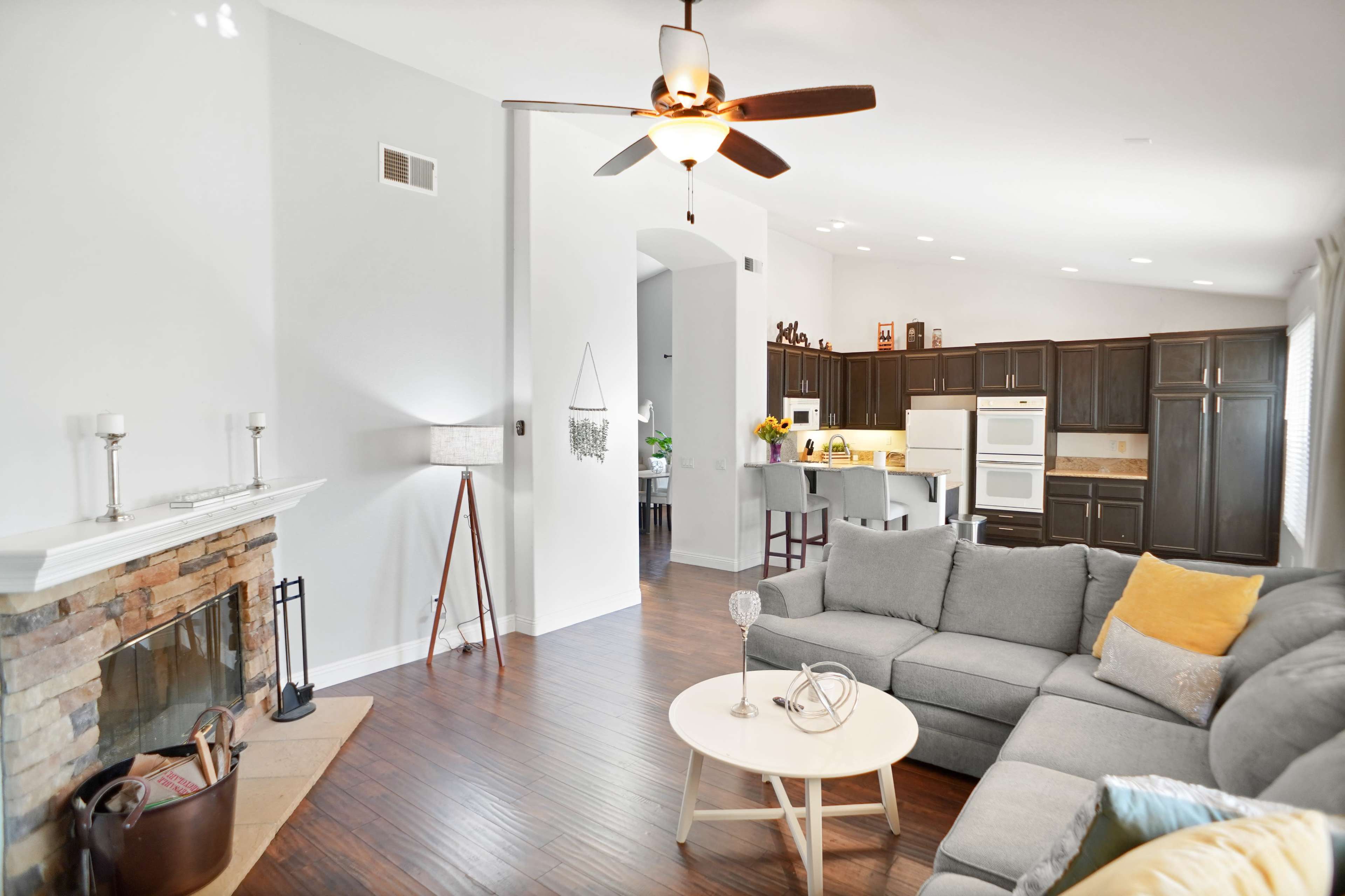 The image shows a modern living room with a gray sectional sofa, a round coffee table, and a stone fireplace, leading to an open kitchen with dark cabinets in the background.