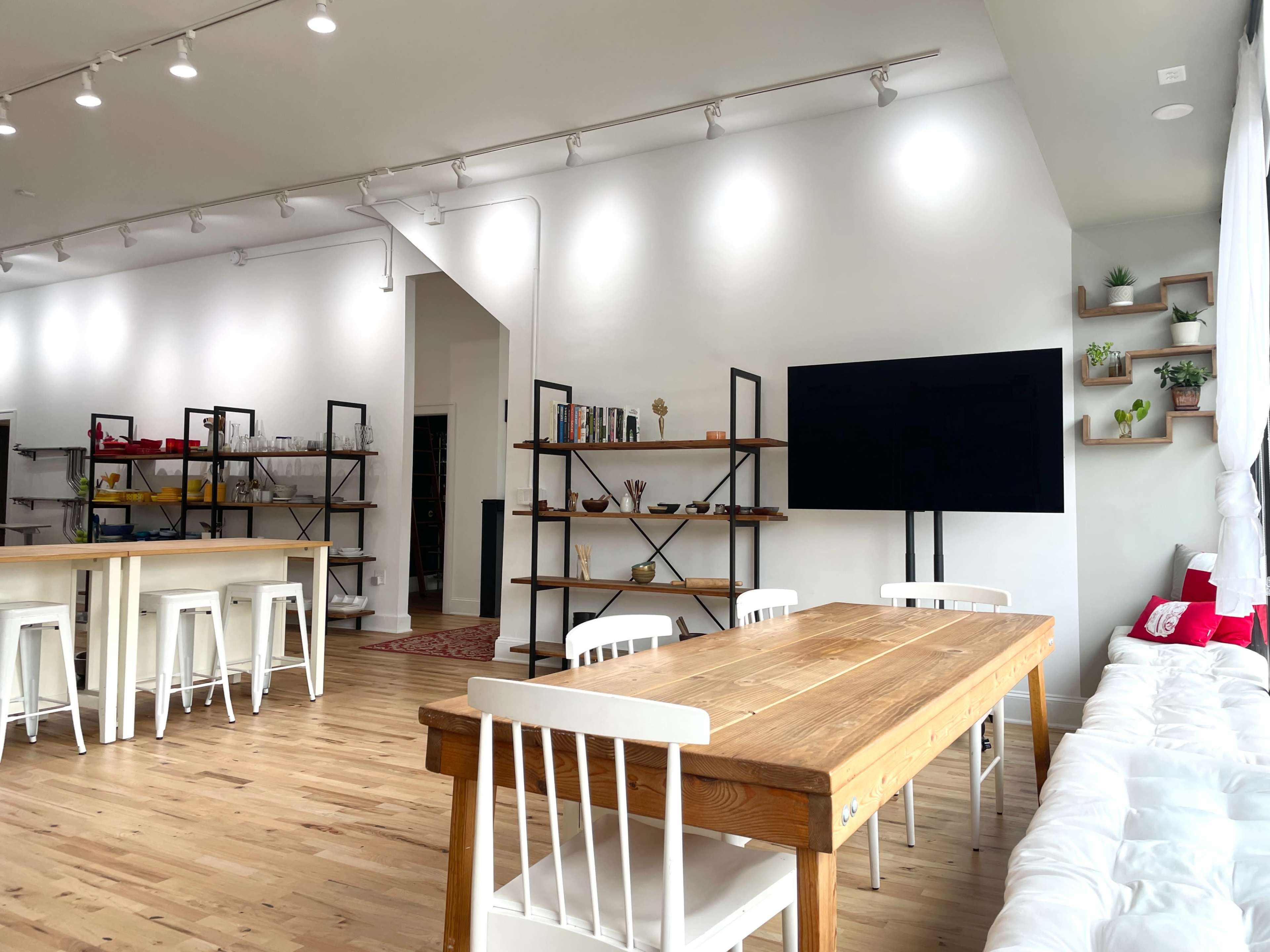 The image shows a modern, open-concept room featuring a wooden dining table, white chairs, shelving units with various items, and a large television mounted on the wall.