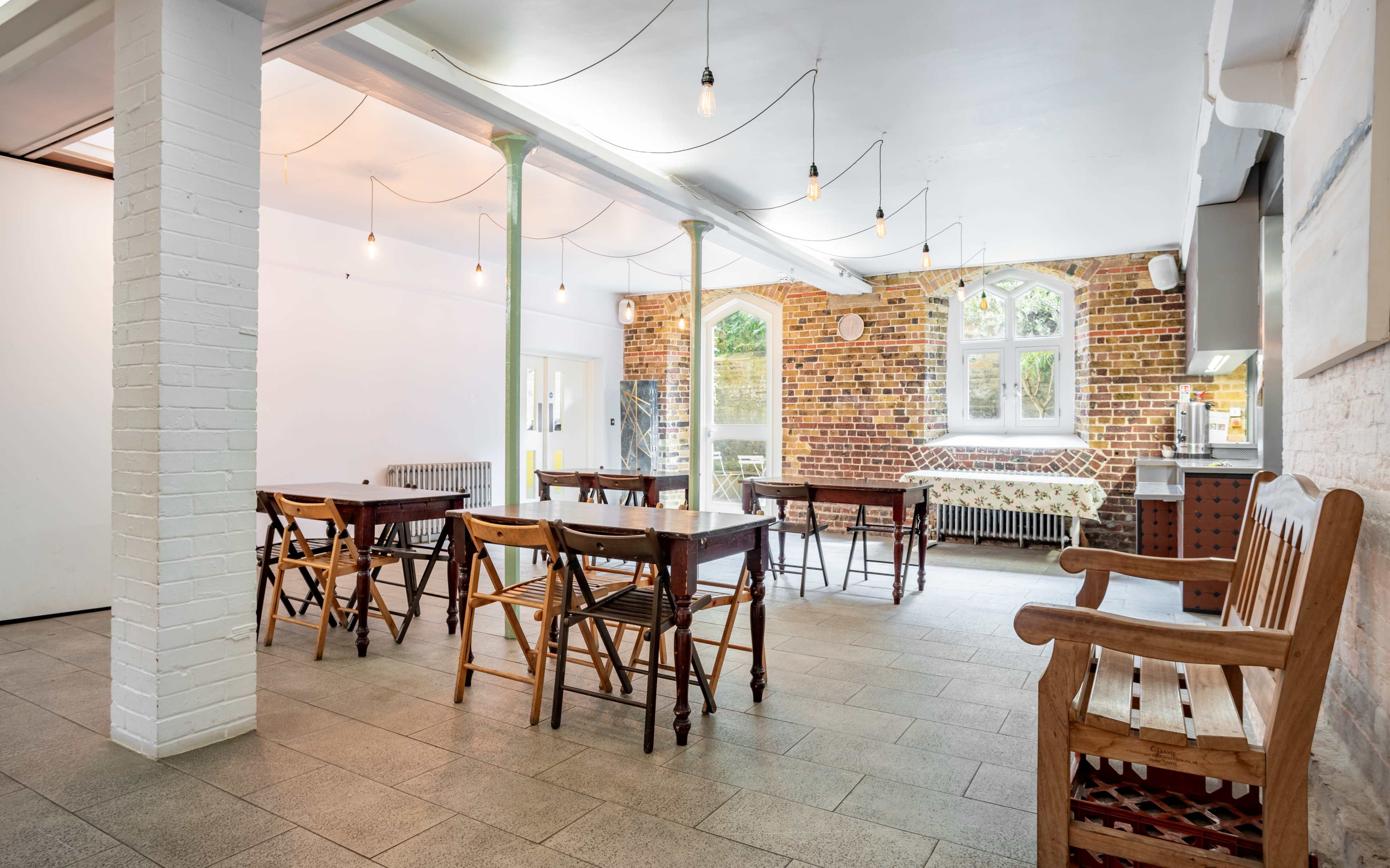 A spacious dining area with wooden tables and chairs, brick walls, and large windows, illuminated by hanging light bulbs.