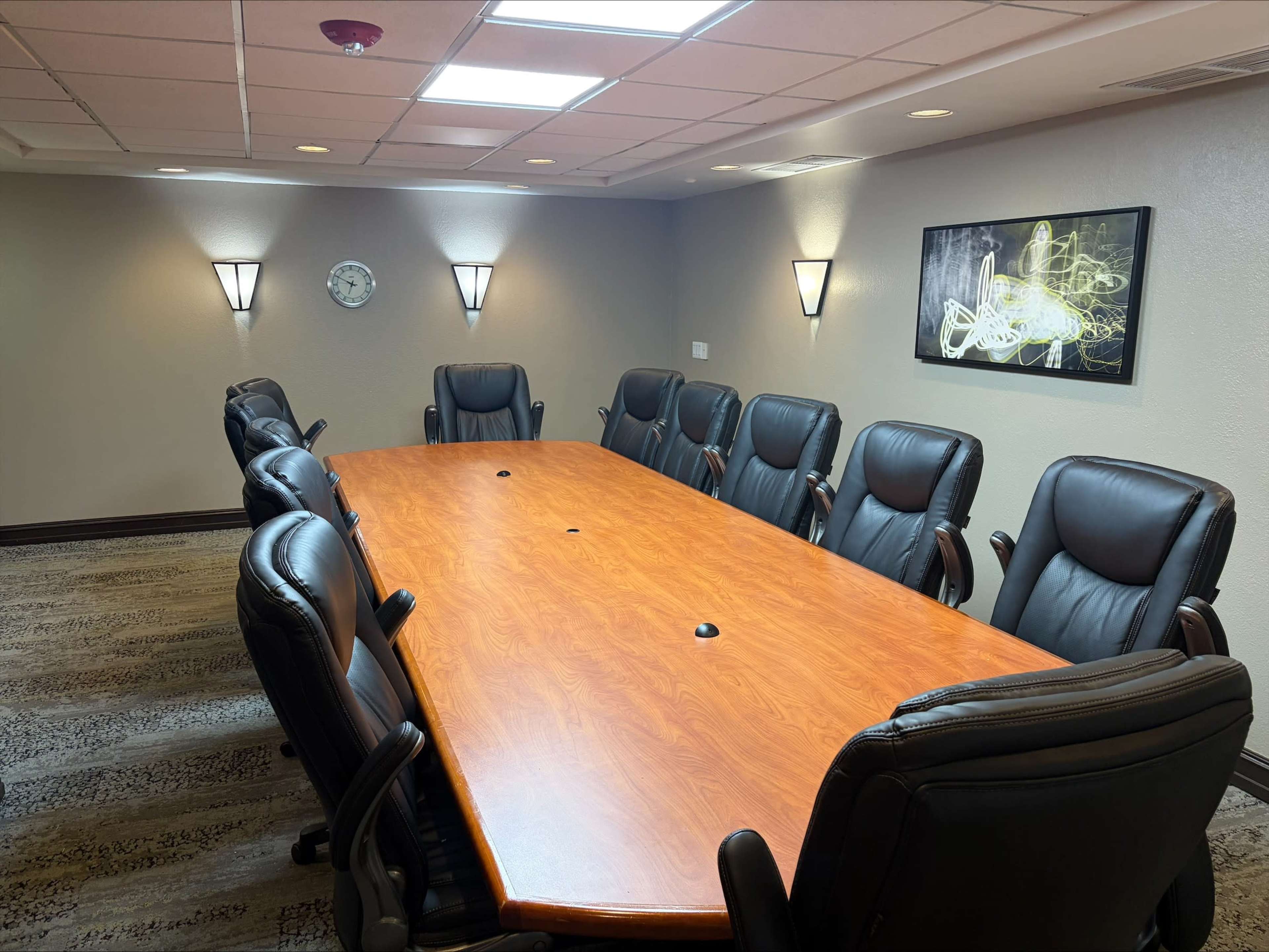 The image shows a conference room with a large wooden table surrounded by ten black leather chairs, illuminated by wall sconces and overhead lights.
