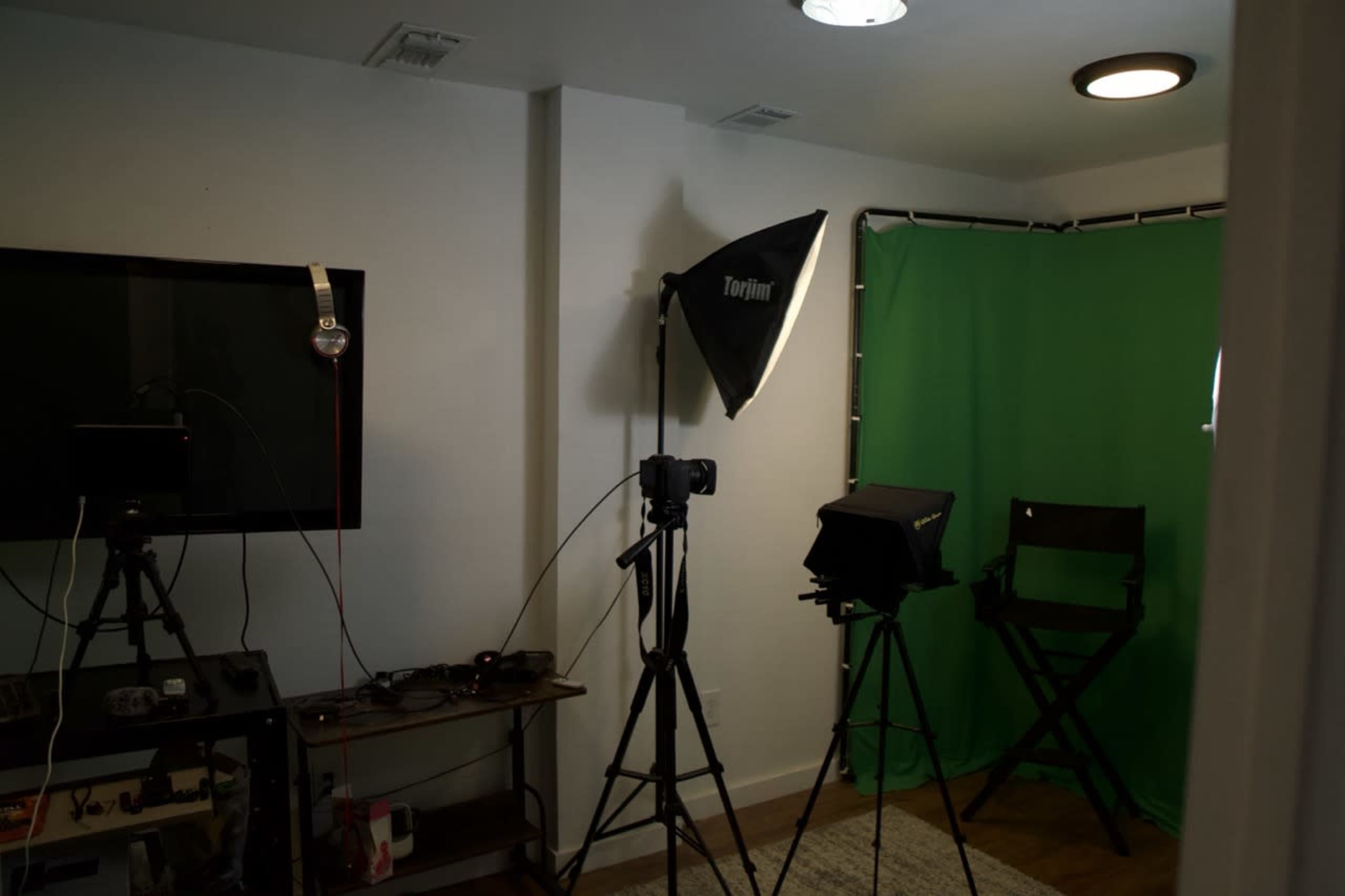 The image shows a small studio setup with a green backdrop, a camera on a tripod, studio lighting, and a director's chair.