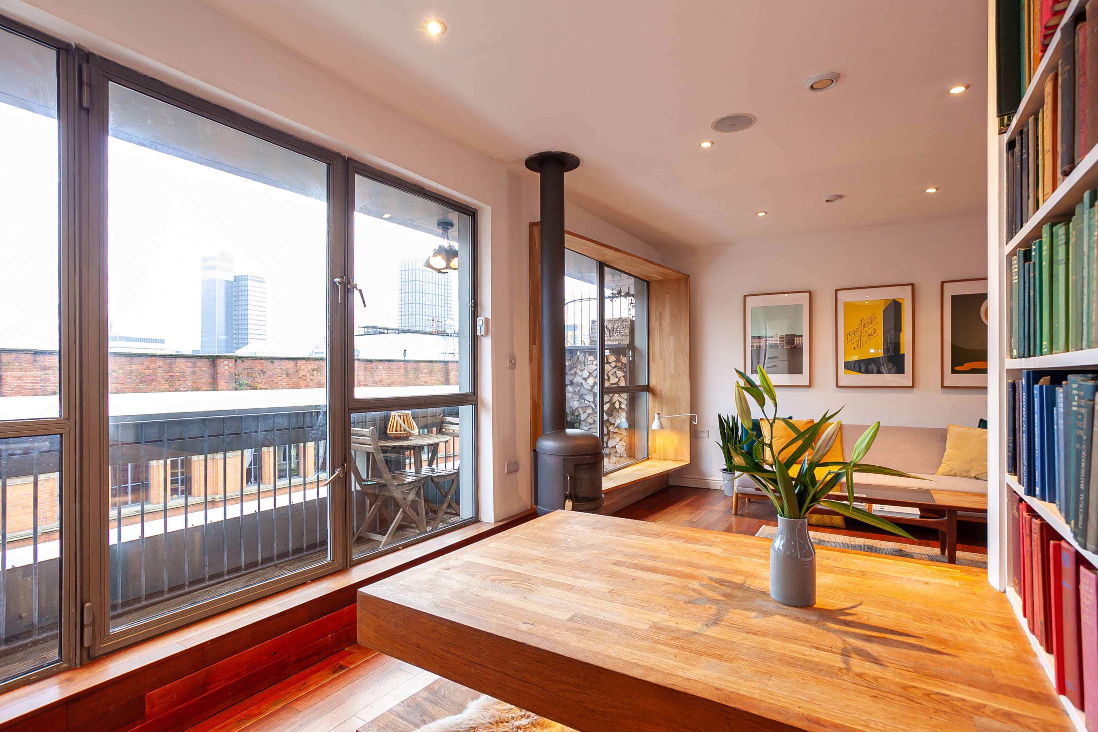 A modern living space with large windows, featuring a wooden table, a cozy seating area, and a small stove, overlooking a cityscape.