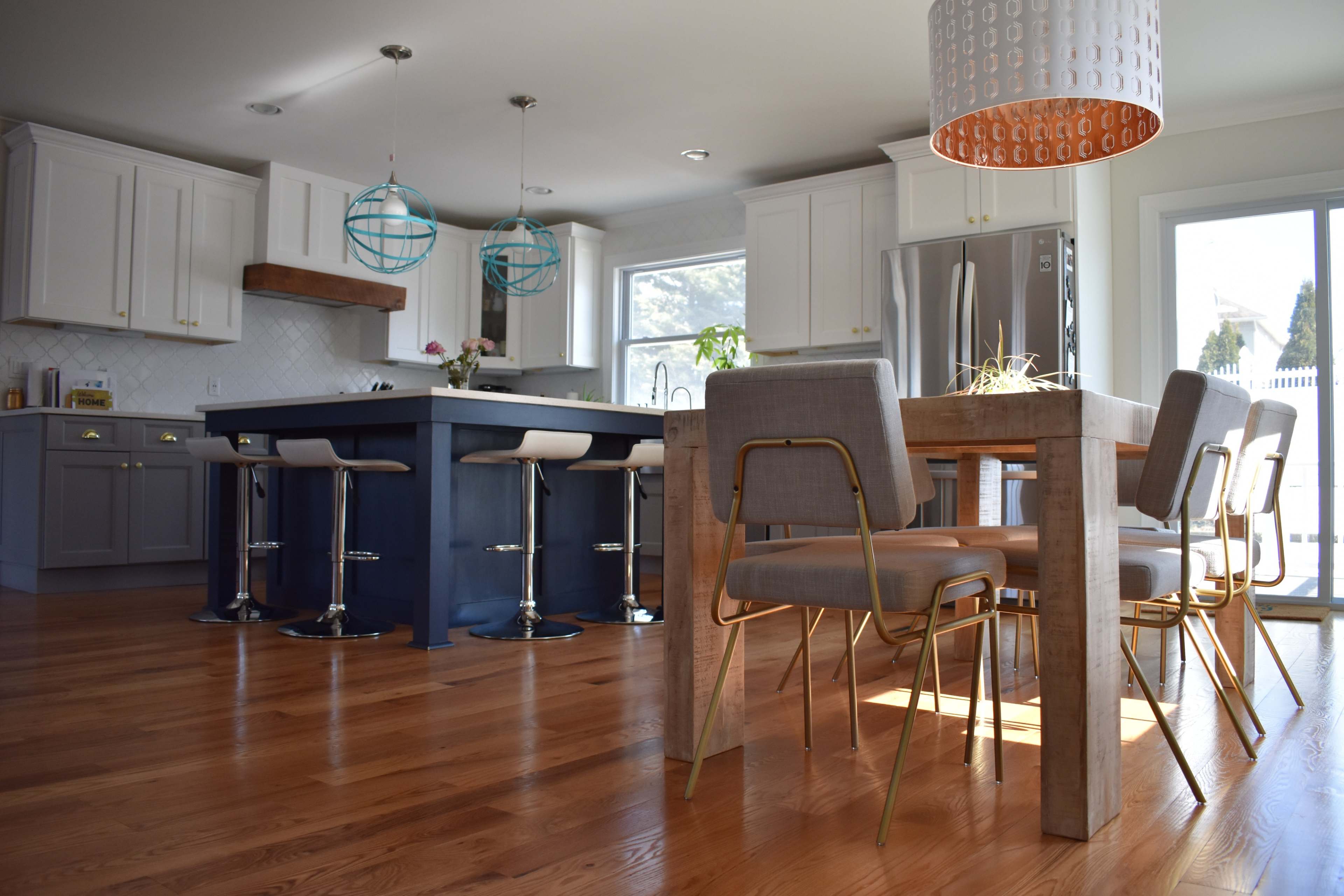 A modern kitchen features a central island with bar stools, a dining table with metallic chairs, and large windows allowing natural light to fill the space.