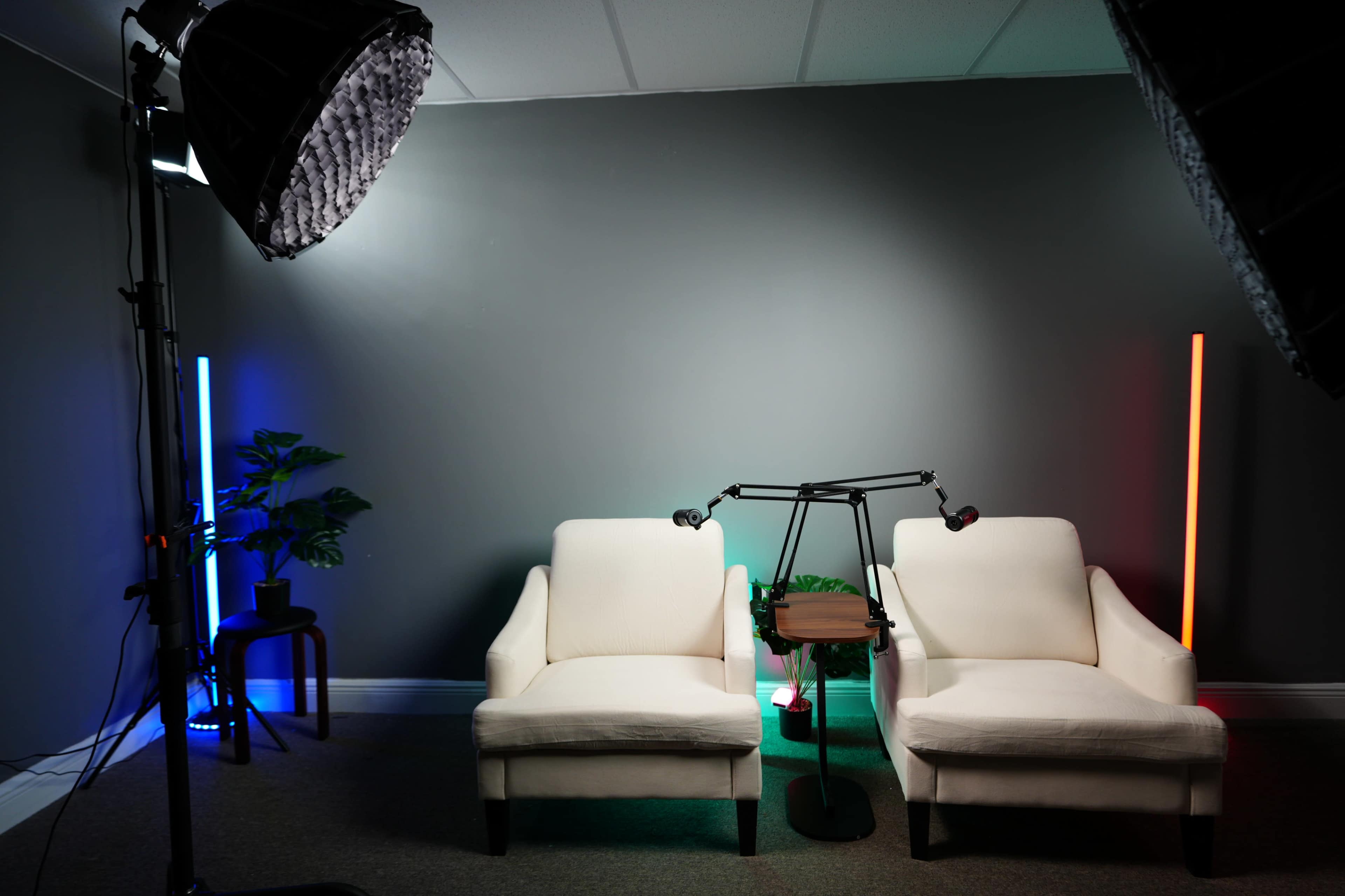 The image shows a cozy set designed for a video recording, featuring two white armchairs facing a small wooden table, with colorful lighting and a potted plant nearby.
